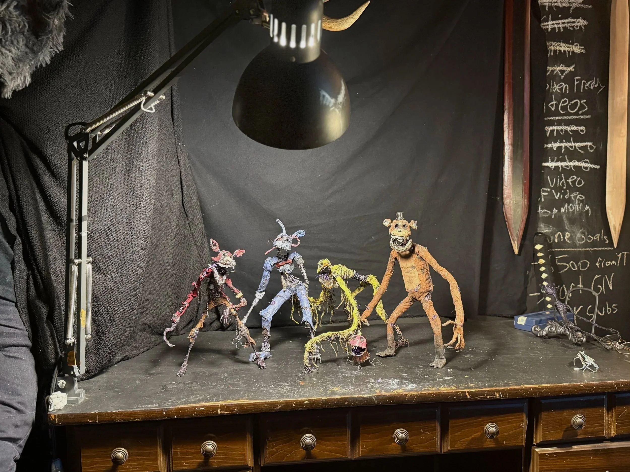 Clay or puppet figures resembling animals and monsters are displayed on a black tabletop against a black backdrop, with a chalkboard and canoe paddles hanging on the wall to the right.