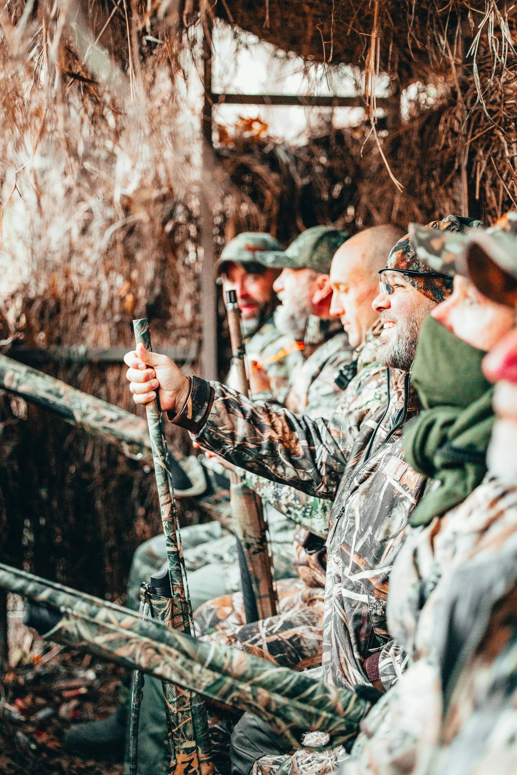 A group of hunters sitting together in camouflage clothing, sitting inside a camouflage hunting blind, with their rifles resting on their laps.