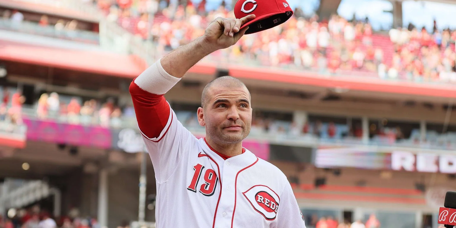 A man in a Cincinnati Reds baseball jersey with the number 19, holding a Cincinnati Reds cap, at a stadium.