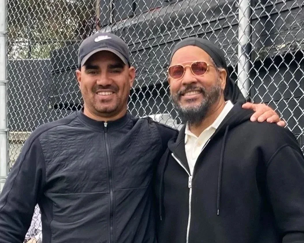 Cody Fitzsimmons standing close together with Manny Ramirez, smiling, with a chain-link fence in the background. One man is wearing a black Nike cap and black jacket, the other man is wearing sunglasses, a bandana, a black hoodie, and a white shirt.