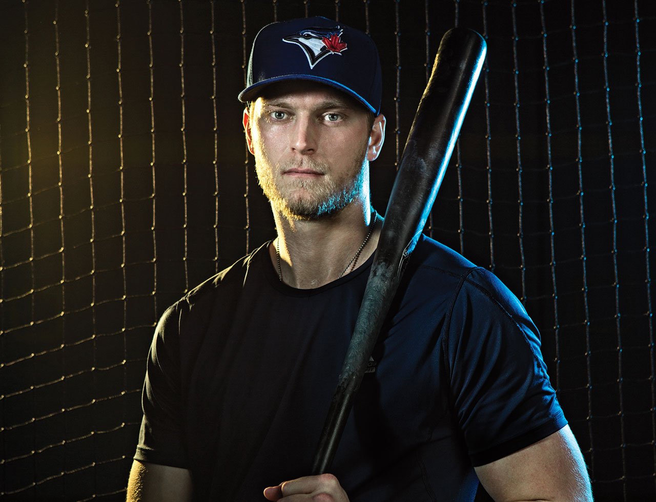 A young man in a Toronto Blue Jays baseball cap holding a baseball bat, standing in front of a net.