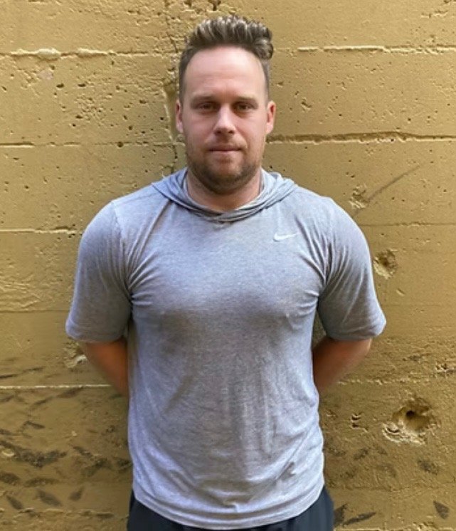 Josh Campbell Strength & Conditioning Coach against a textured yellowish-brown wall, wearing a light gray Nike T-shirt, with his hands behind his back, looking directly into the camera.