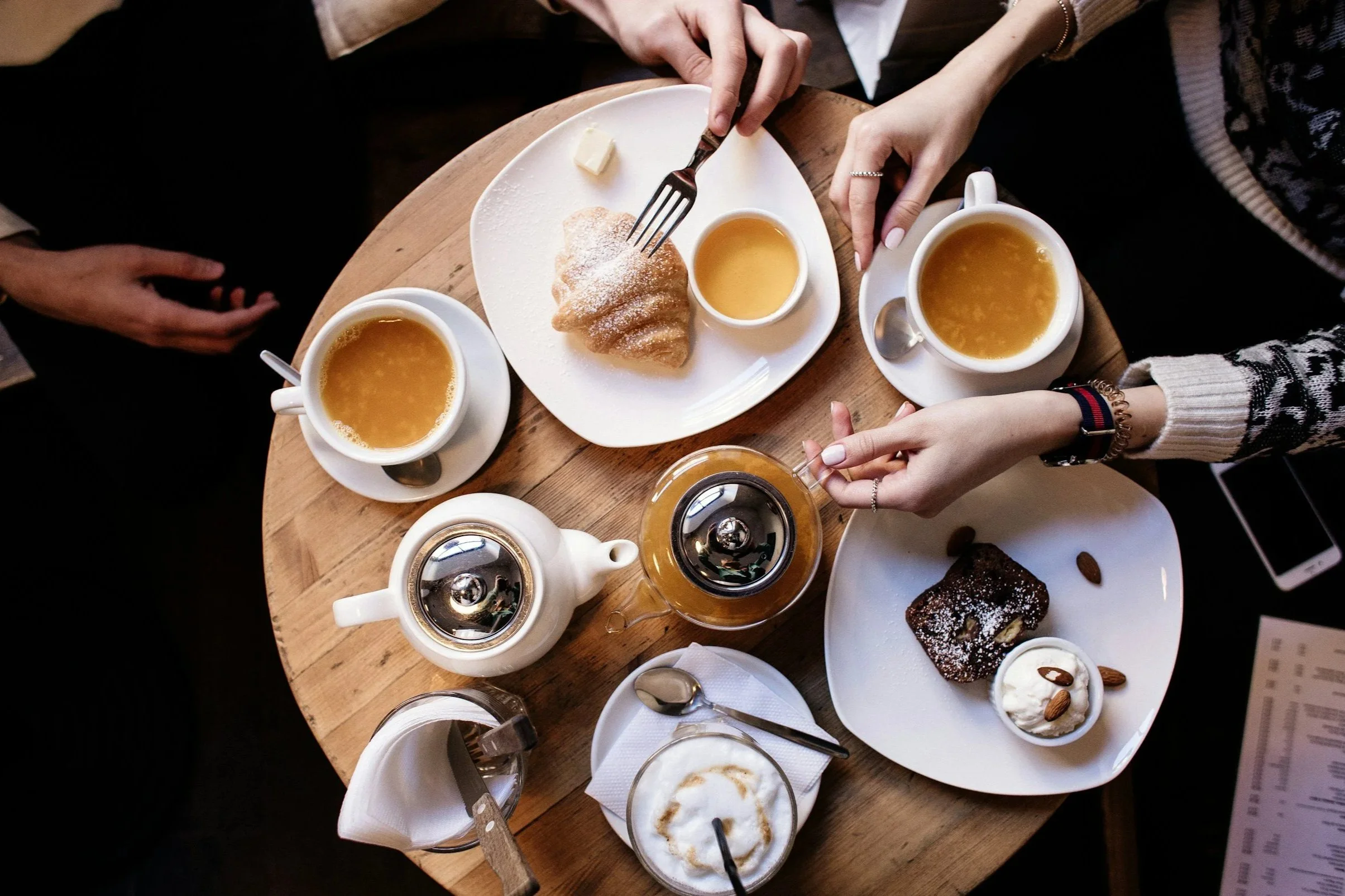 Top-down view of a round wooden table set for afternoon tea with two cups of tea, a teapot, a glass of cappuccino, a plate with a croissant, a plate with a piece of chocolate cake, cream, almonds, and a small jar of honey, with two people reaching for food.