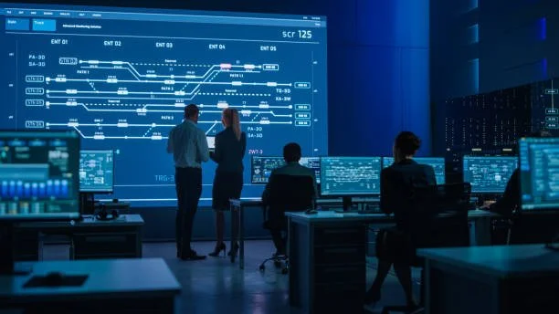 Control room with multiple monitors showing train schedules and operations, with four people working and one person standing and discussing near a large display.