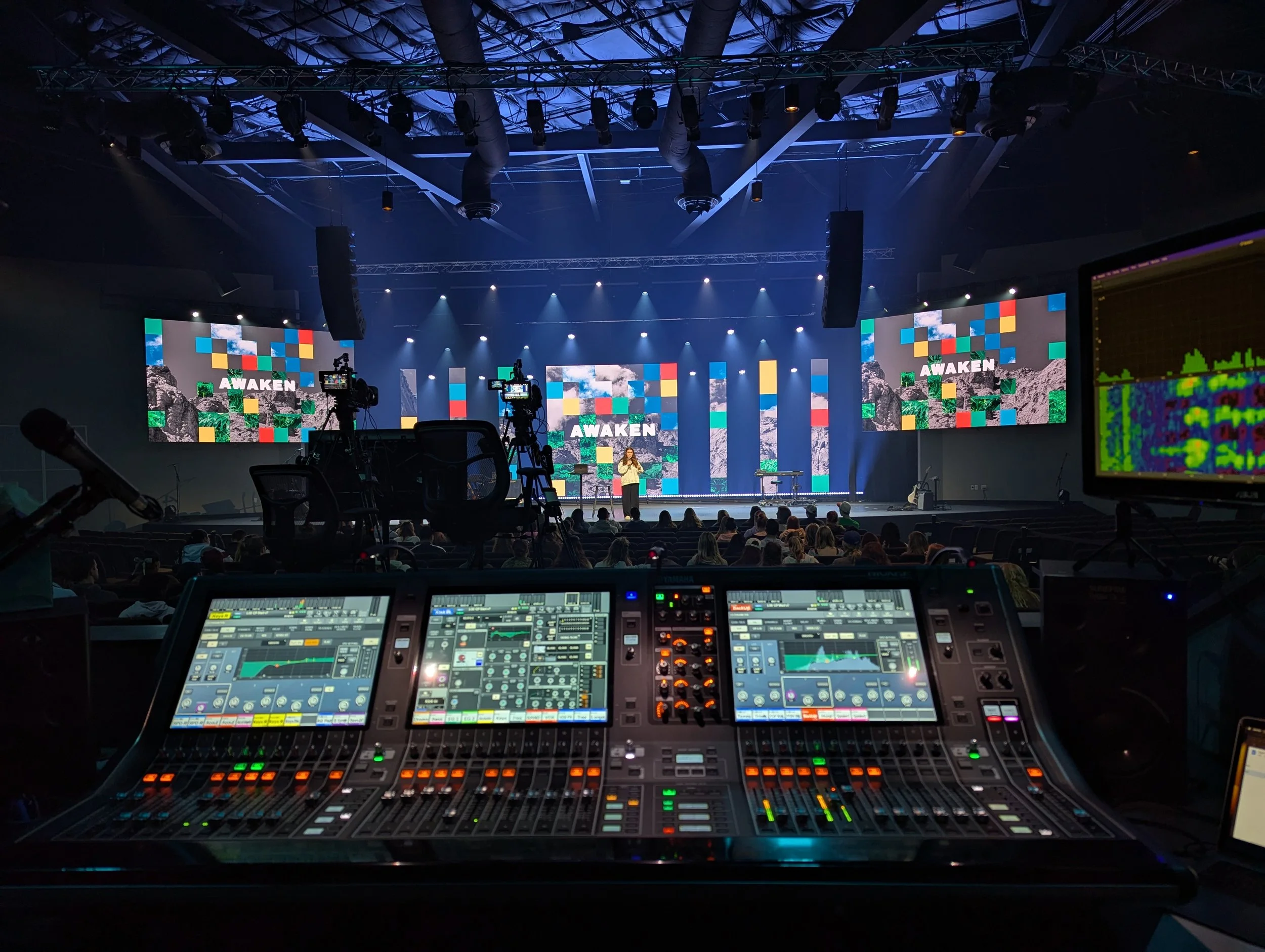 The image shows a stage with large screens displaying the word 'AWAKEN' along with colorful graphics and a speaker giving a presentation. In the foreground, there is sound and lighting equipment, including a mixer board and cameras, while the audience can be seen seated facing the stage.