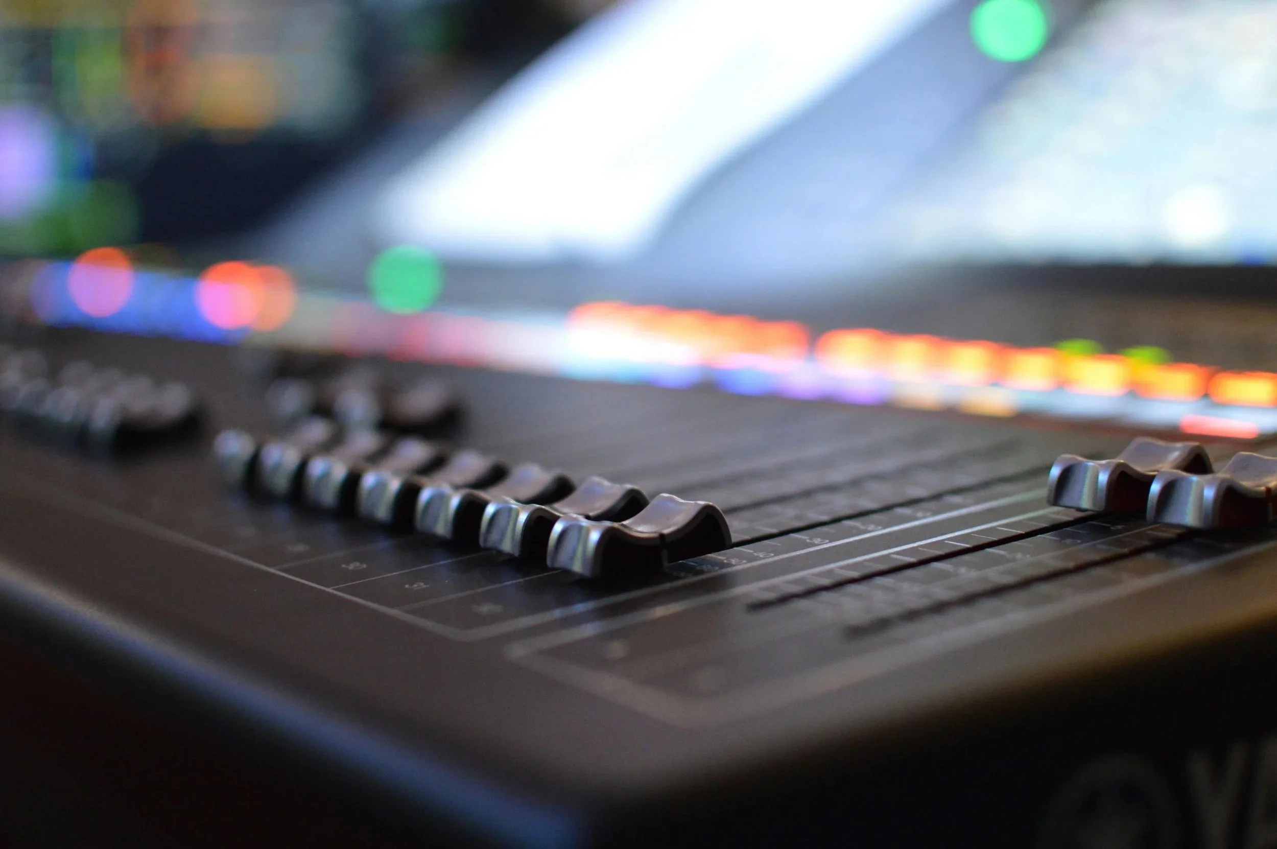 Close-up of a music mixing console with colorful illuminated sliders and controls, out of focus in the background.