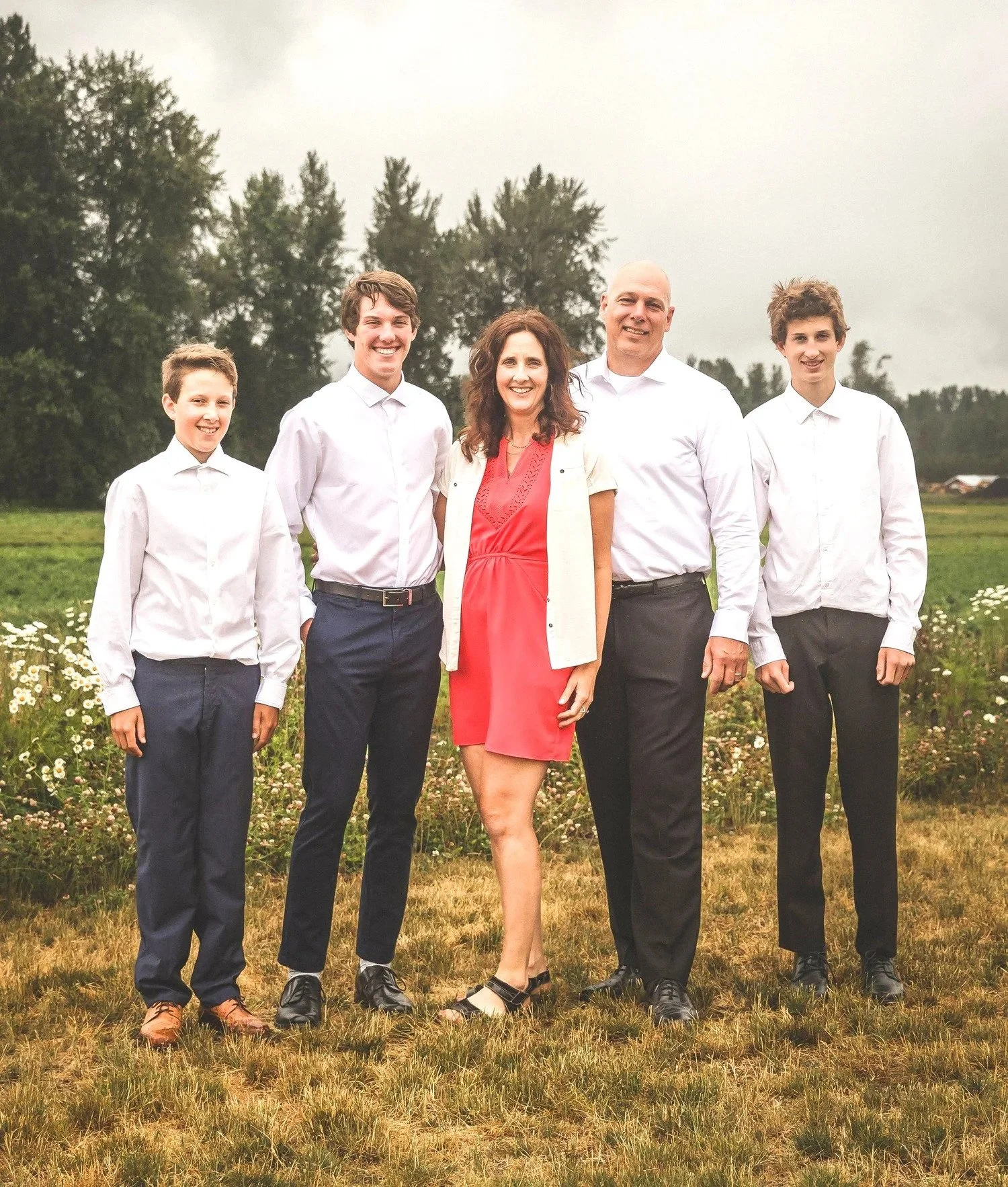 A family of five standing outdoors on grass with trees and overcast sky in background, dressed in semi-formal attire, smiling at the camera.