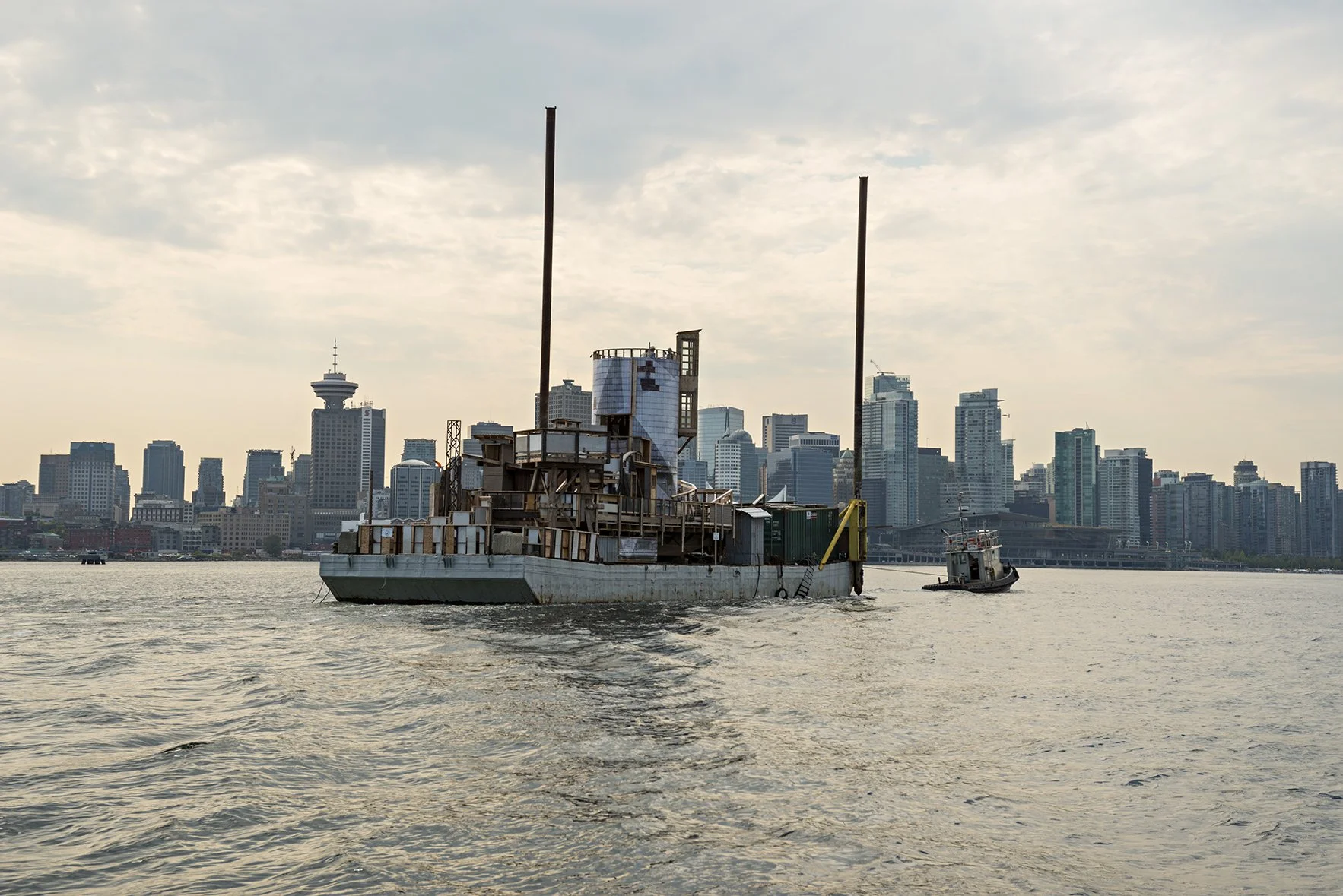 A weathered barge floating on a body of water with a city skyline in the background, including skyscrapers and a prominent tower.