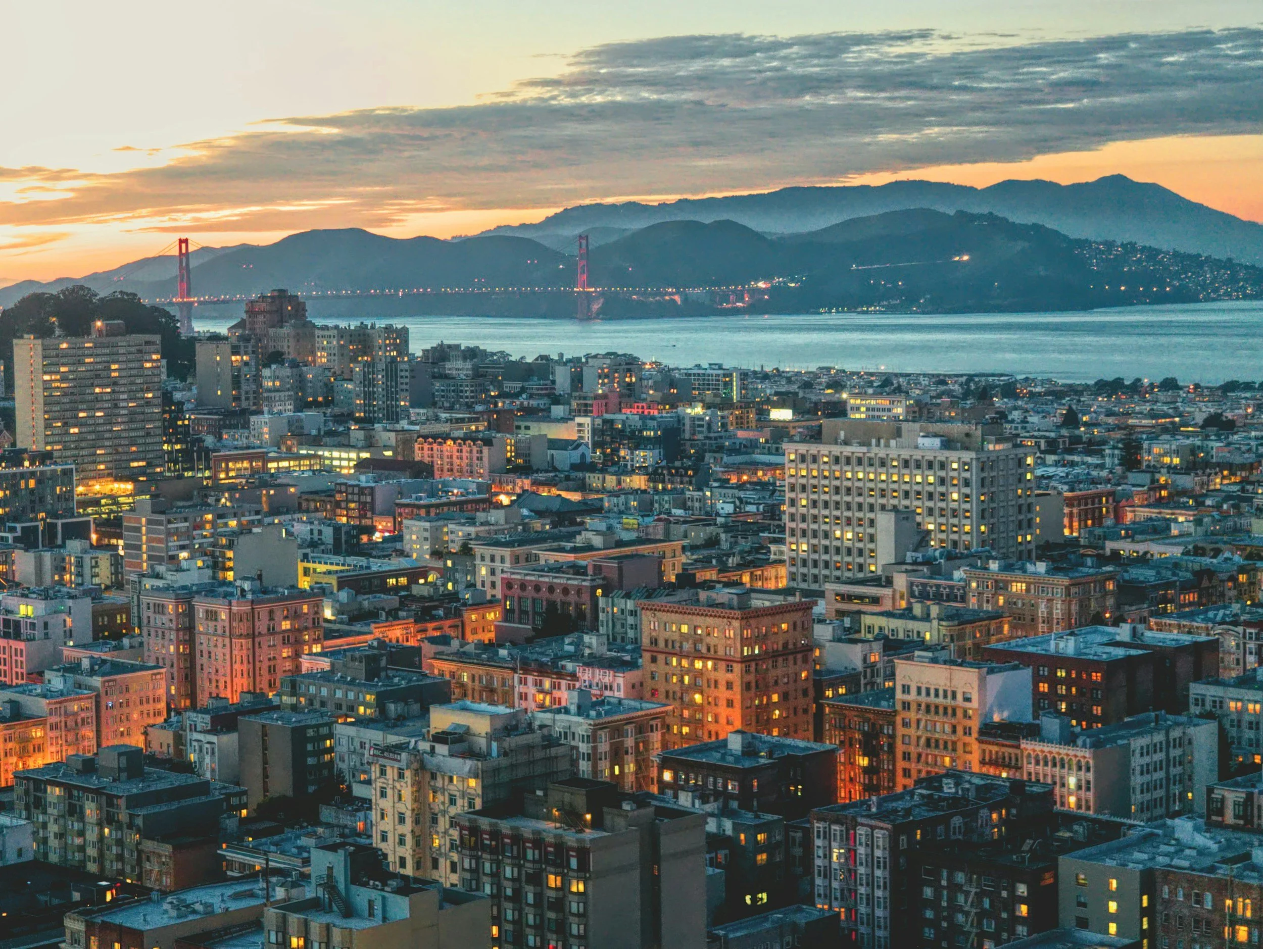 San Francisco cityscape at sunset with the Golden Gate Bridge and hills in the background, and buildings with lit windows in the foreground.