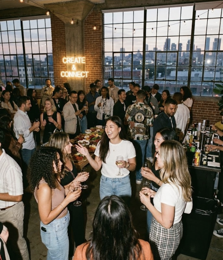 People socializing at a party or networking event held in an industrial-style venue with large windows, exposed brick walls, and string lights. A neon sign on the wall reads 'CREATE & CONNECT'.
