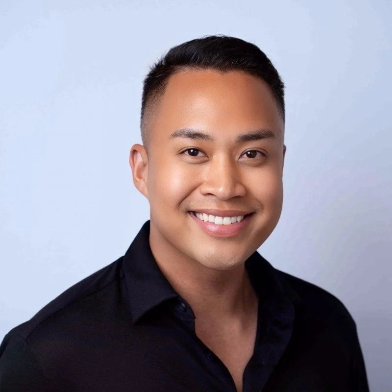 Smiling young man with short black hair wearing a black collared shirt, against a light gray background.