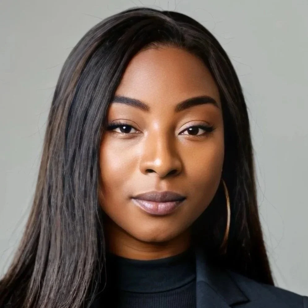 Close-up portrait of a young woman with straight, long dark hair, light makeup, and hoop earrings, wearing a dark top against a neutral background.