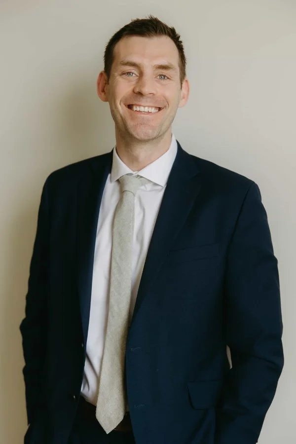 Russell Lytle, smiling in a navy blue suit, white shirt, and light-colored tie, standing against a plain off-white wall.