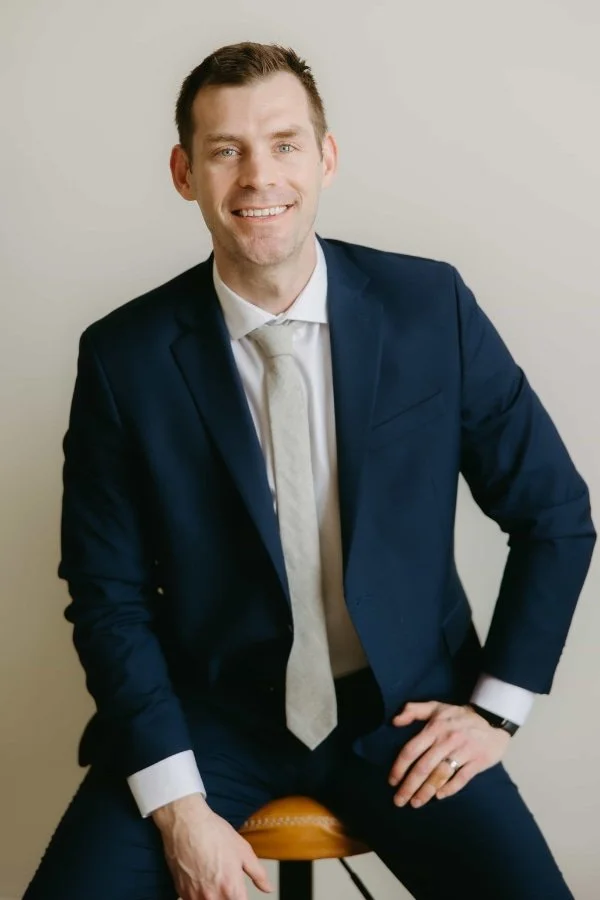 Russell Lytle, head of Lytle Law in a dark blue suit and a white tie, sitting on a stool against a plain light-colored wall, smiling at the camera.
