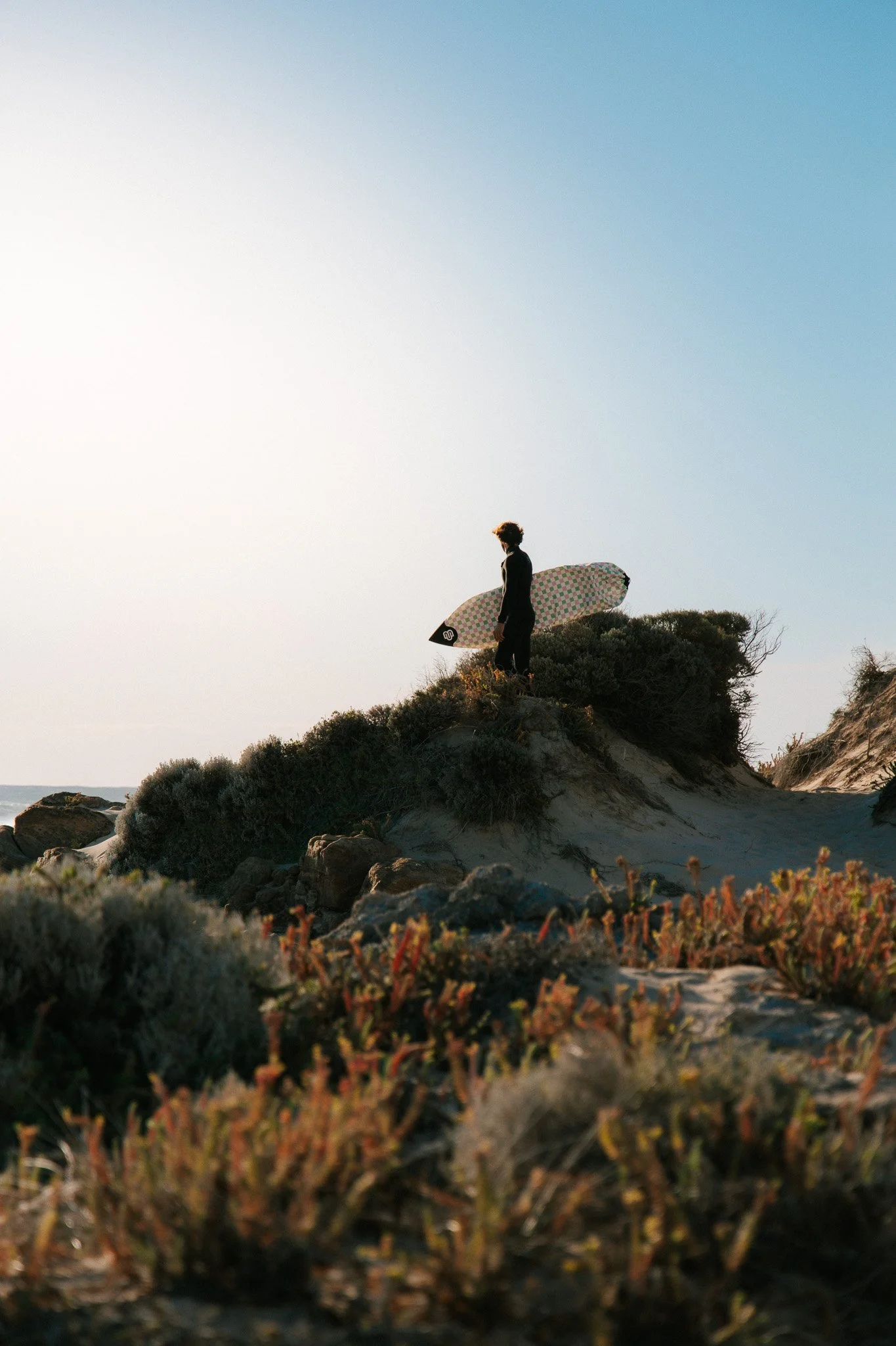 Person standing on a sandy hill holding a surfboard, facing the sky at sunset or sunrise, with coastal vegetation in the foreground.