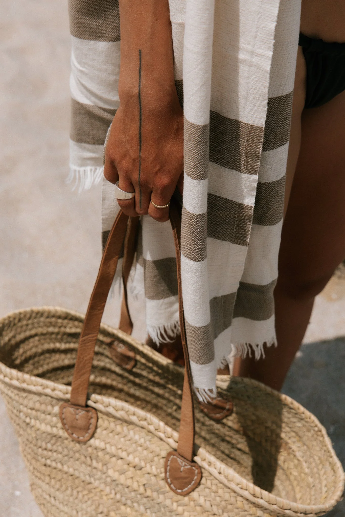A person's hand holding a woven straw bag with leather handles, wearing a checklist ring, in front of a striped towel or blanket.