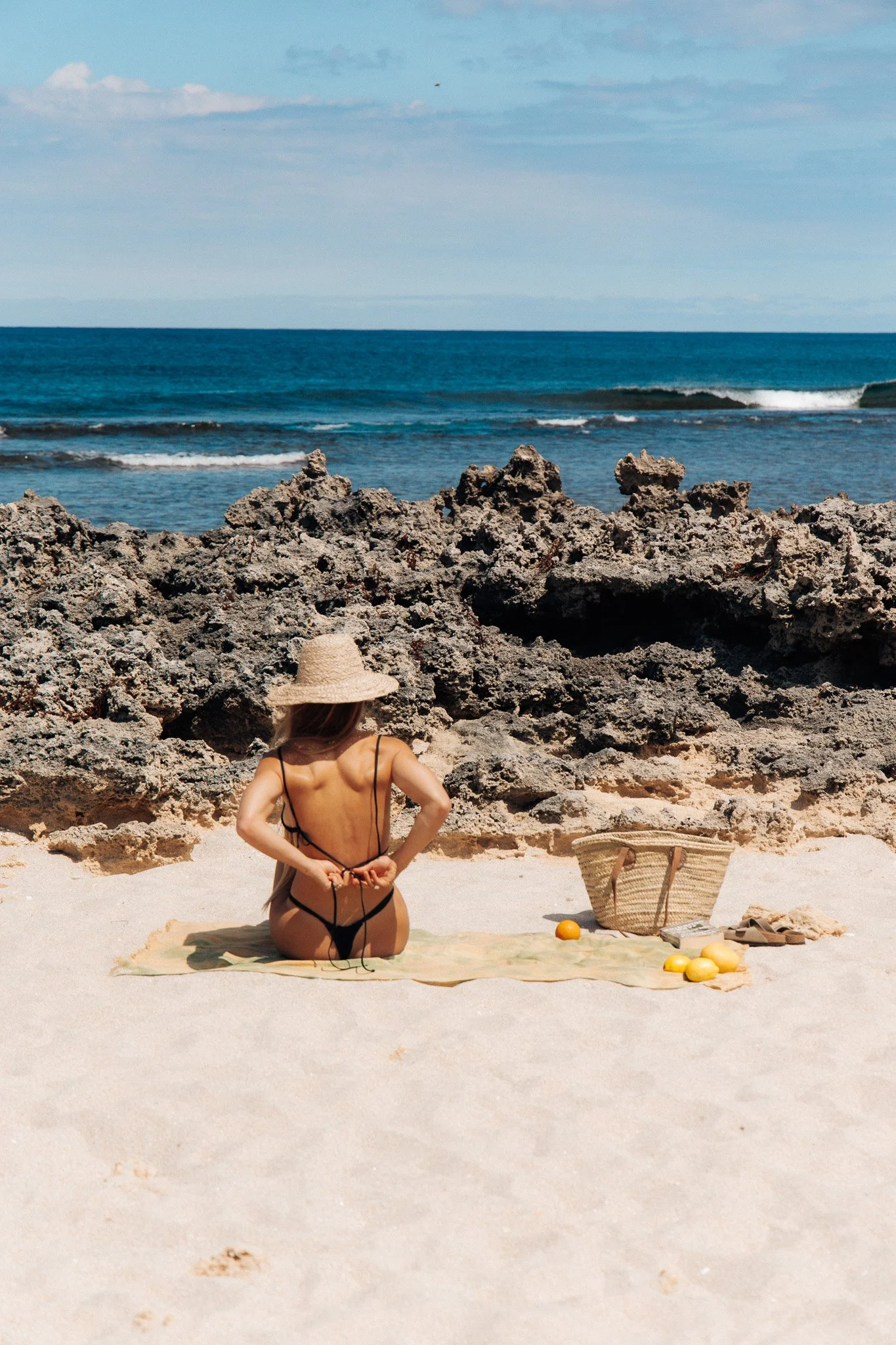 Woman sitting on a towel at the beach with rocks, ocean waves, a basket, a lemon, and oranges nearby.