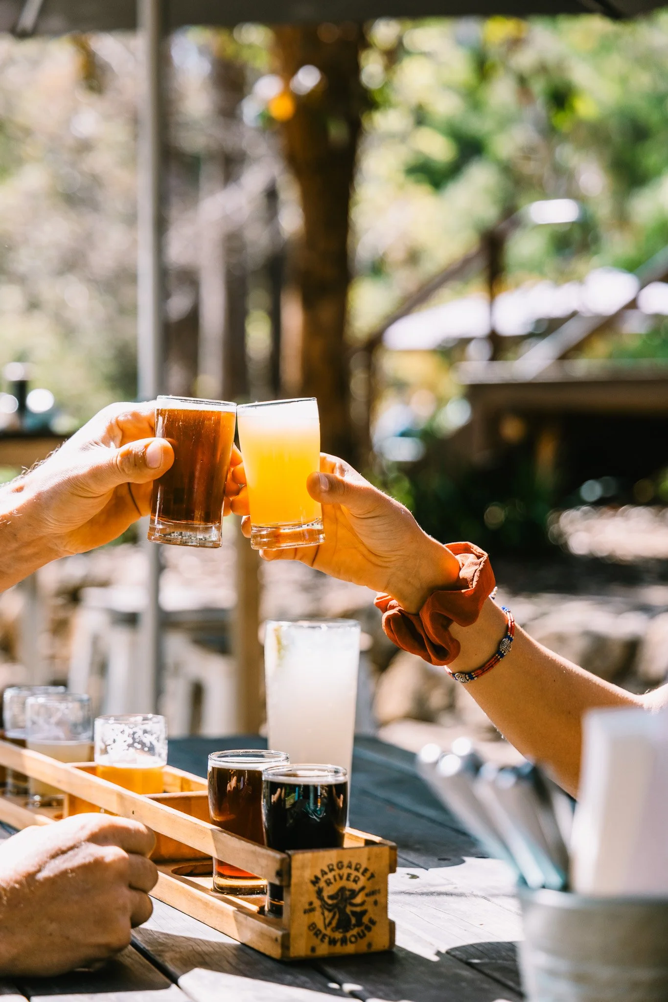 Two people clinking glasses of beer outdoors, with a flight of beers on the table.