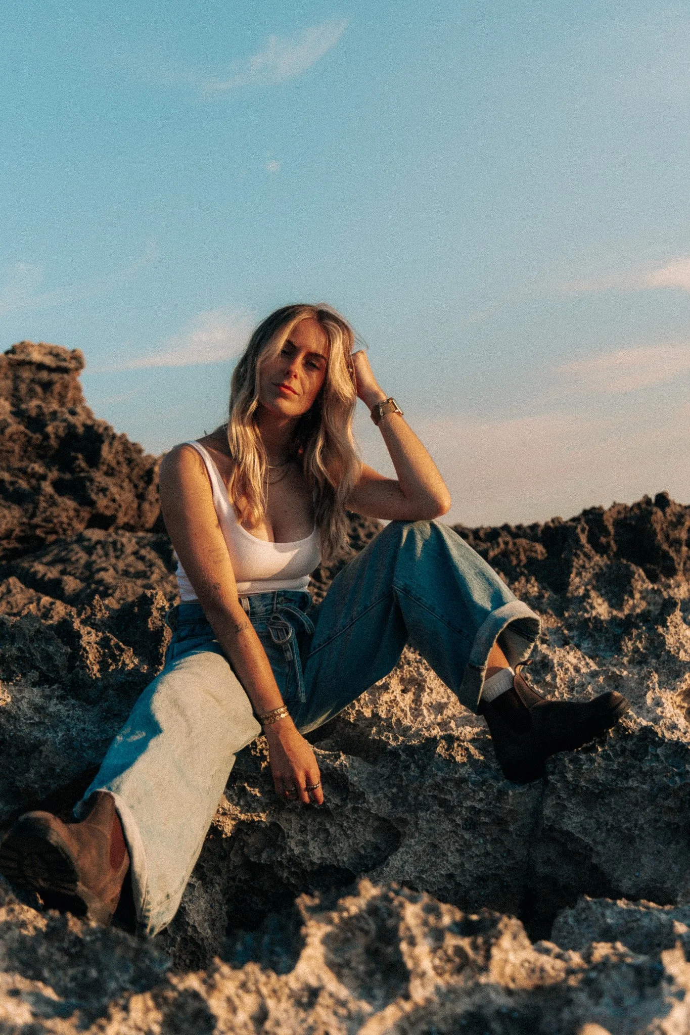 Woman sitting on rocky terrain, wearing a white tank top, jeans, and boots, with a sunset sky in the background.