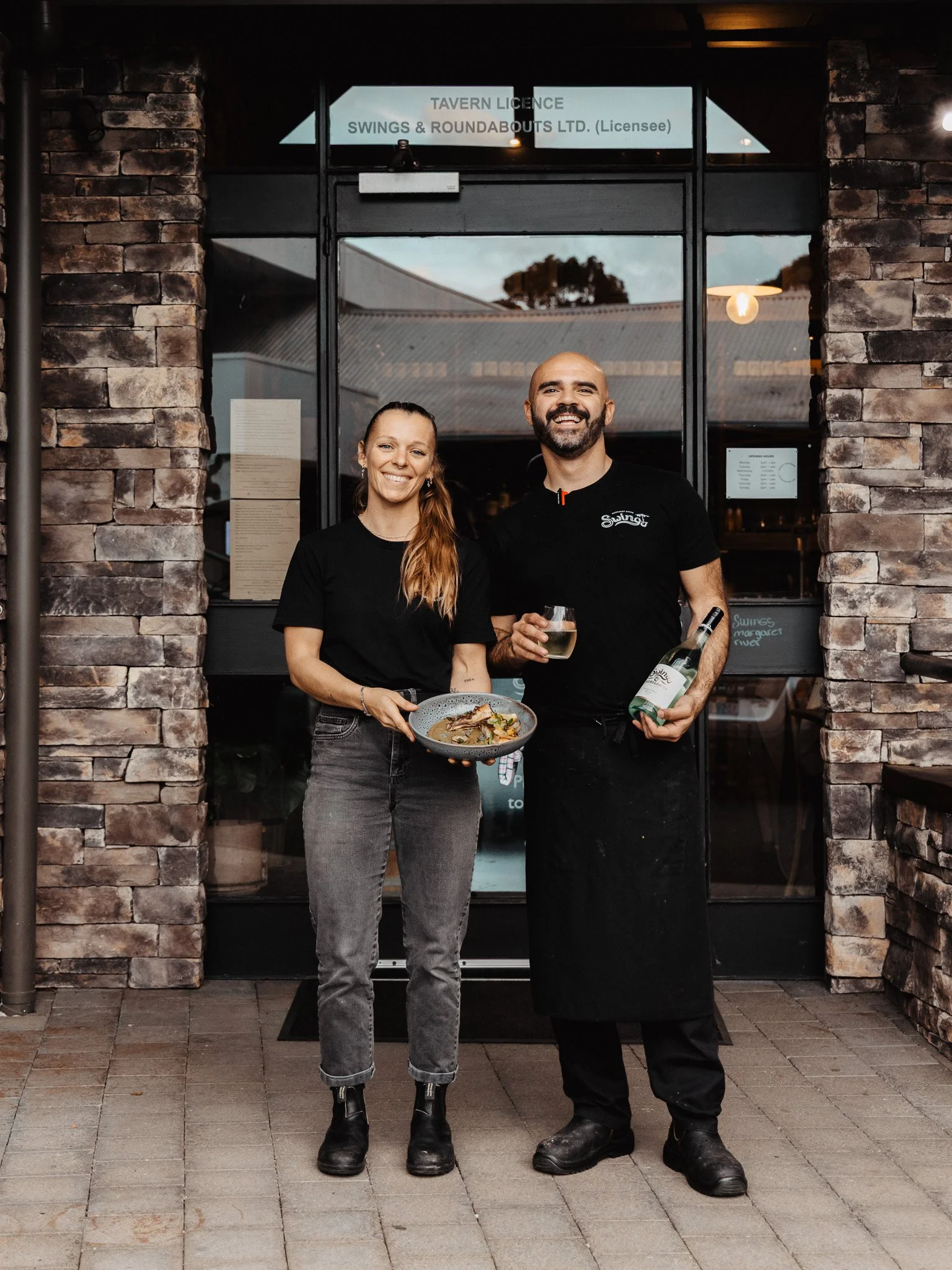 Two smiling restaurant staff members, a woman and a man, standing outside the entrance of a restaurant with brick walls. The woman is holding a plate of food, and the man is holding a glass of wine and a wine bottle.