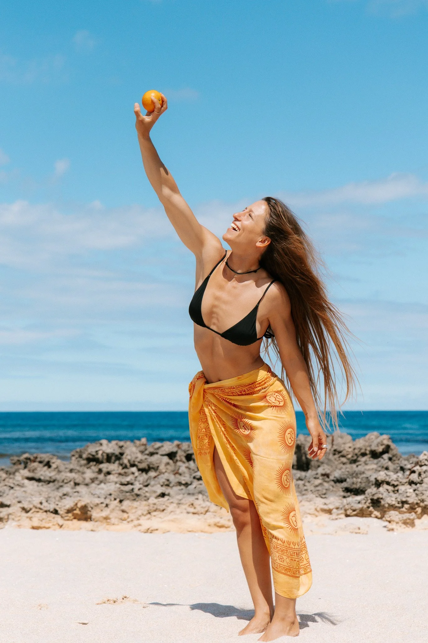 A woman on the beach wearing a black bikini top and a yellow sarong, holding a small orange ball in her raised right hand, smiling and looking up at the sky under a blue sky with some clouds.
