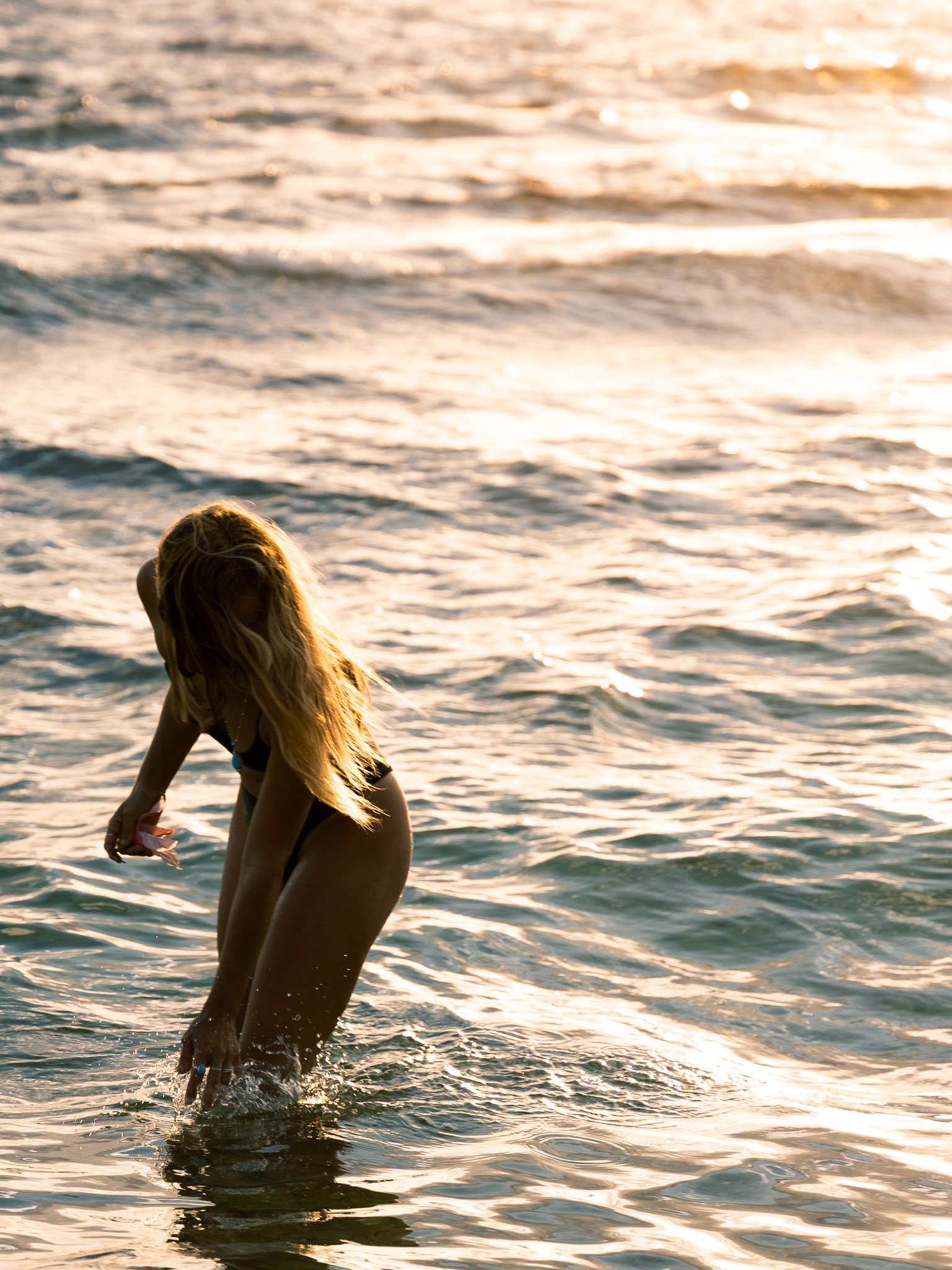 A young woman with long hair standing in the ocean during sunset, adjusting her bikini bottom.