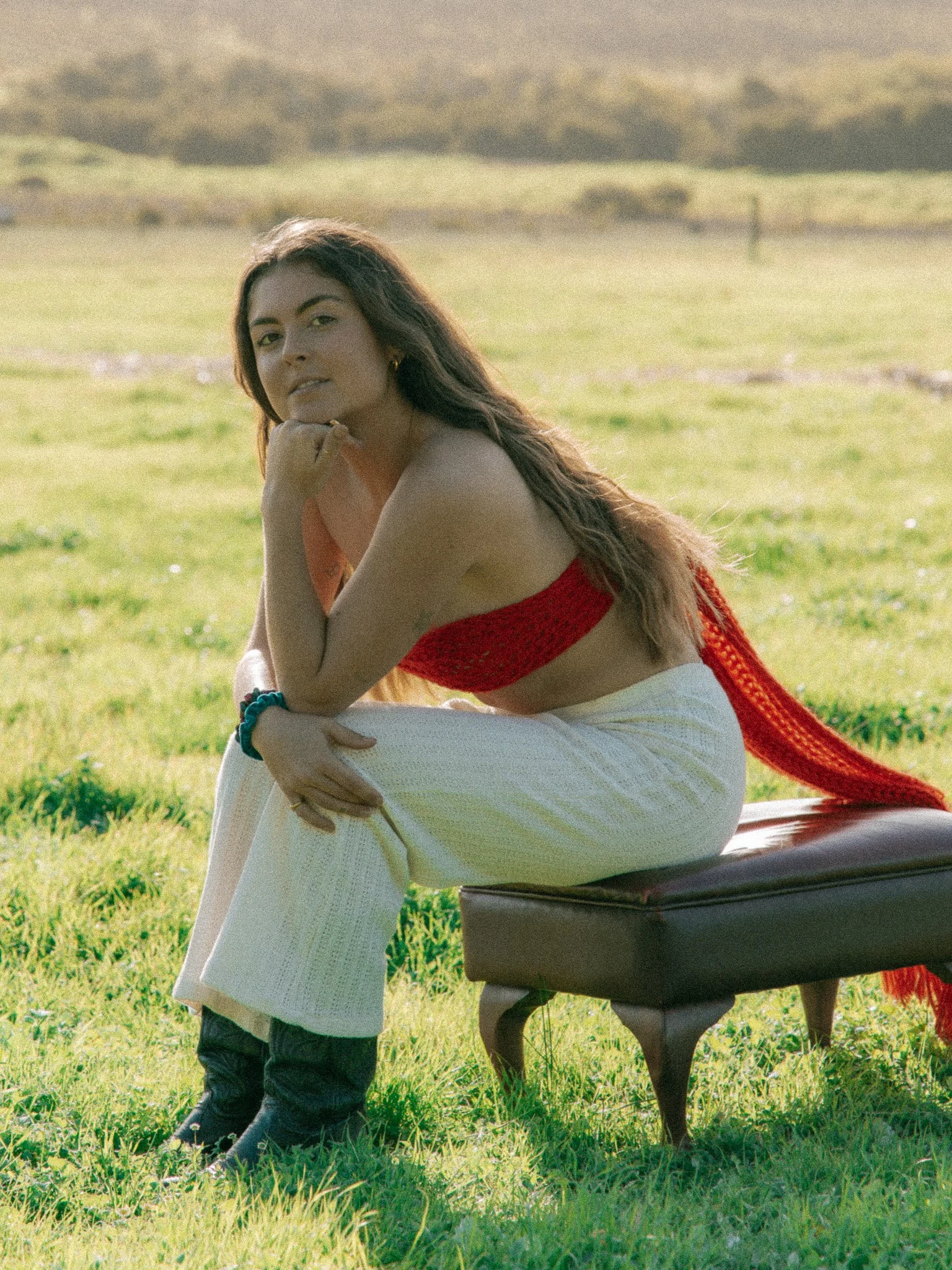 A woman in a red top and white pants sitting on a bench in a grassy outdoor area, with hills in the background.