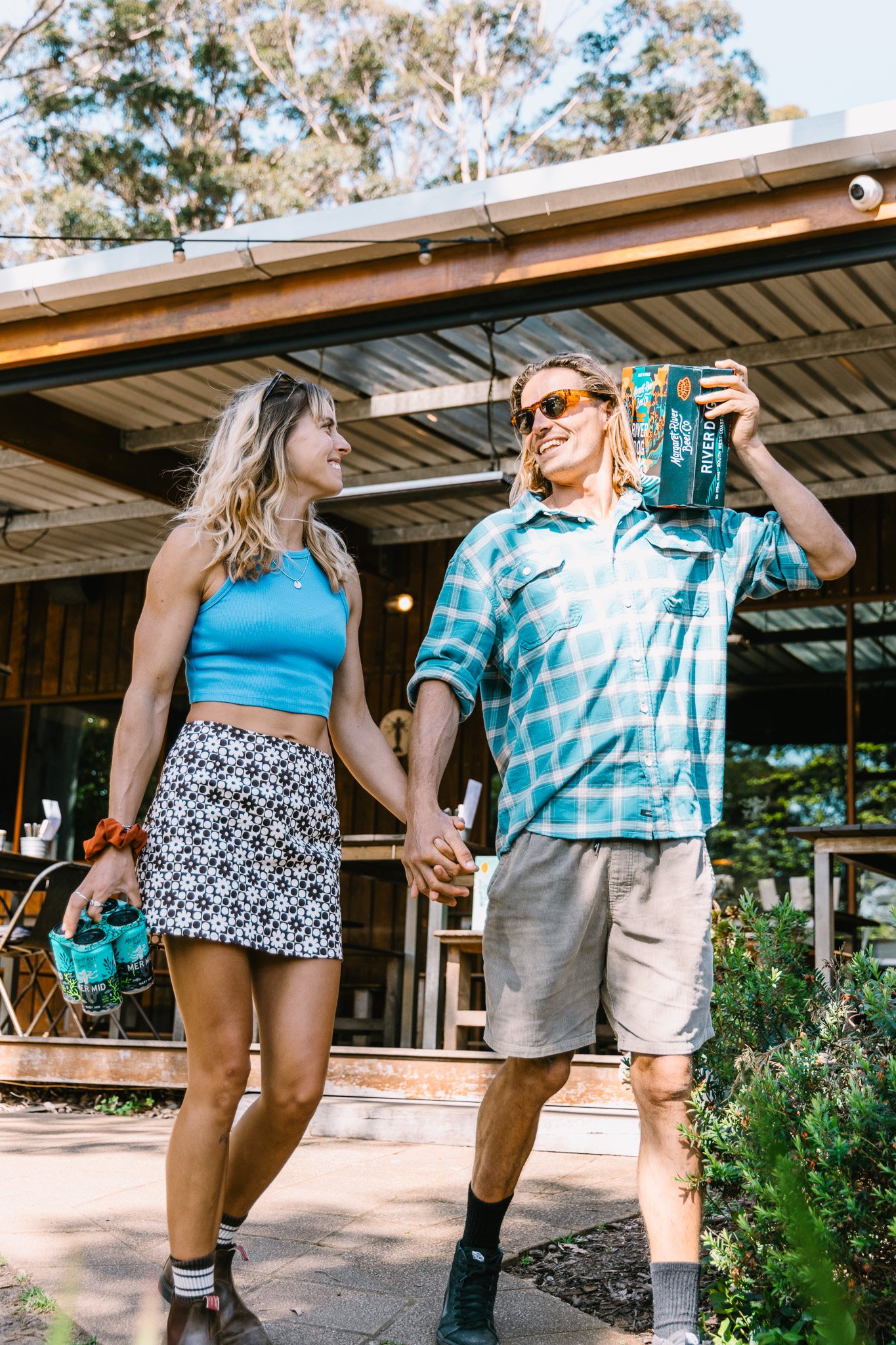 A young woman and man holding hands, smiling, and walking outdoors on a sunny day. The woman is wearing a blue crop top, patterned skirt, and sunglasses, while the man is in a blue checkered shirt, shorts, and sunglasses, carrying a pack of beverages