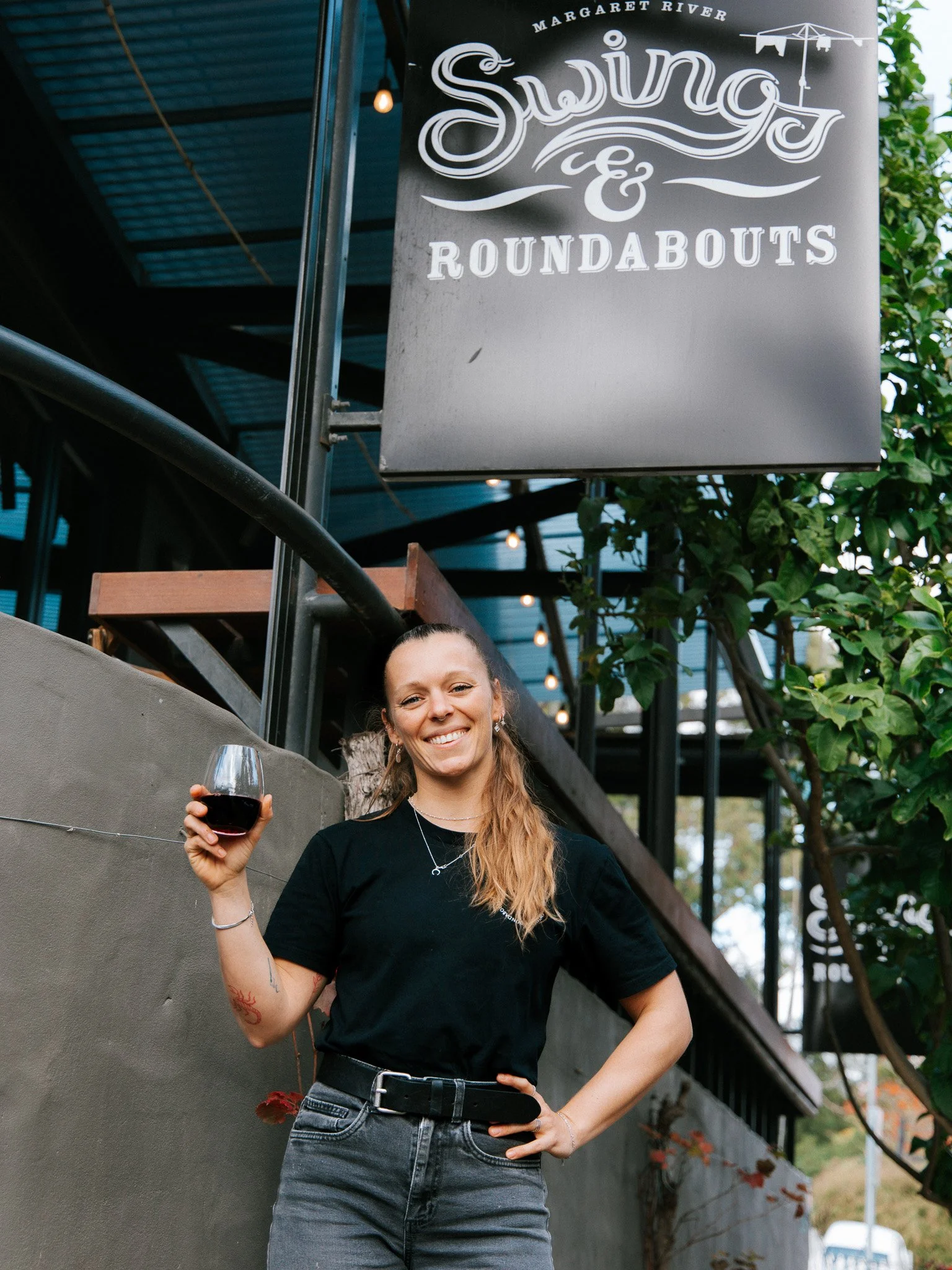 A woman smiling and holding a glass of red wine standing outside at a restaurant or bar with a sign overhead that reads 'Swing & Roundabouts'.