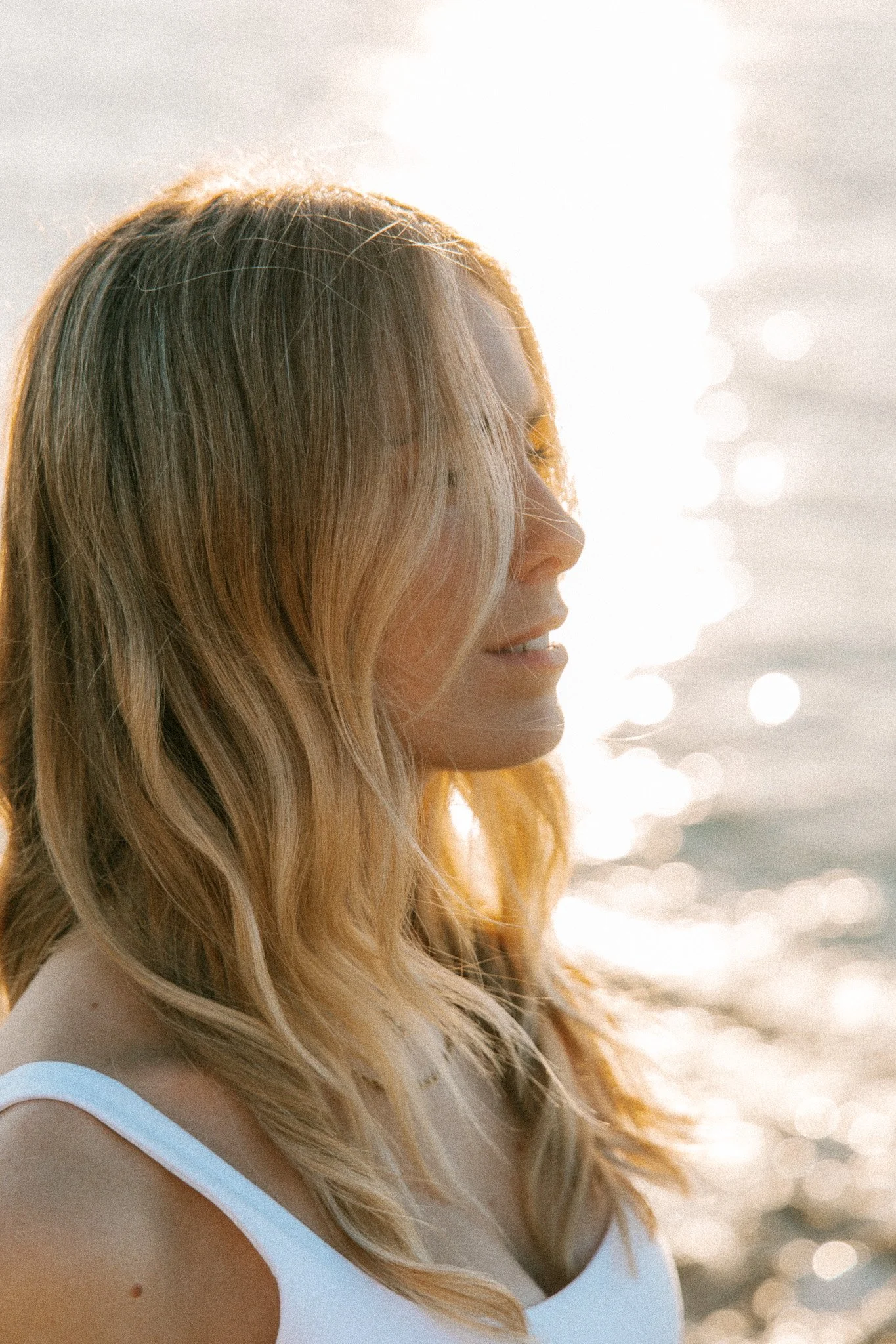 A woman with long, wavy blonde hair is standing near water, smiling softly, with sunlight reflecting off the water in the background.