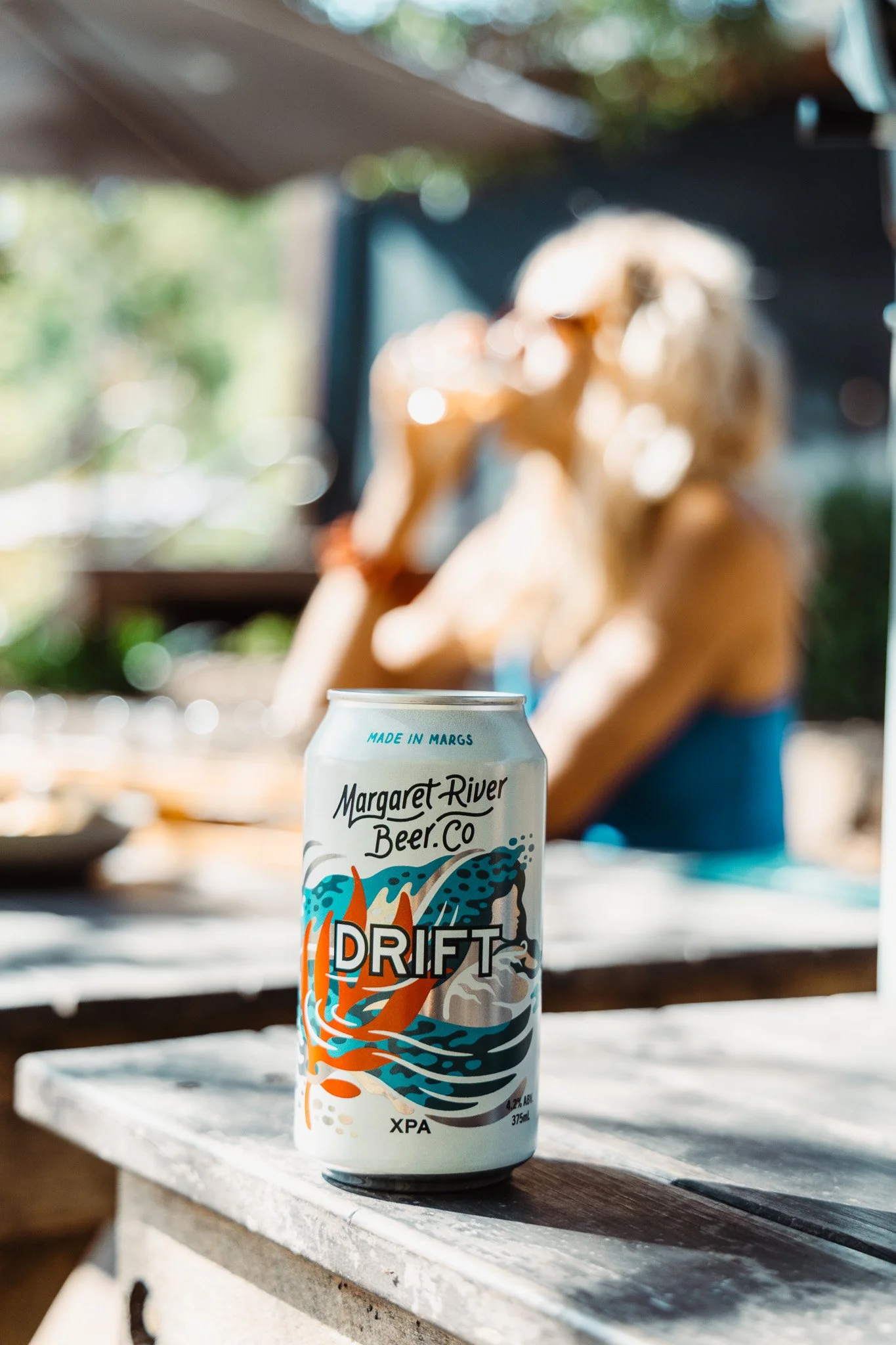A can of Margaret River Beer Co Drift beer on a wooden table with a woman in a blue dress drinking in the background, blurred outdoor setting.
