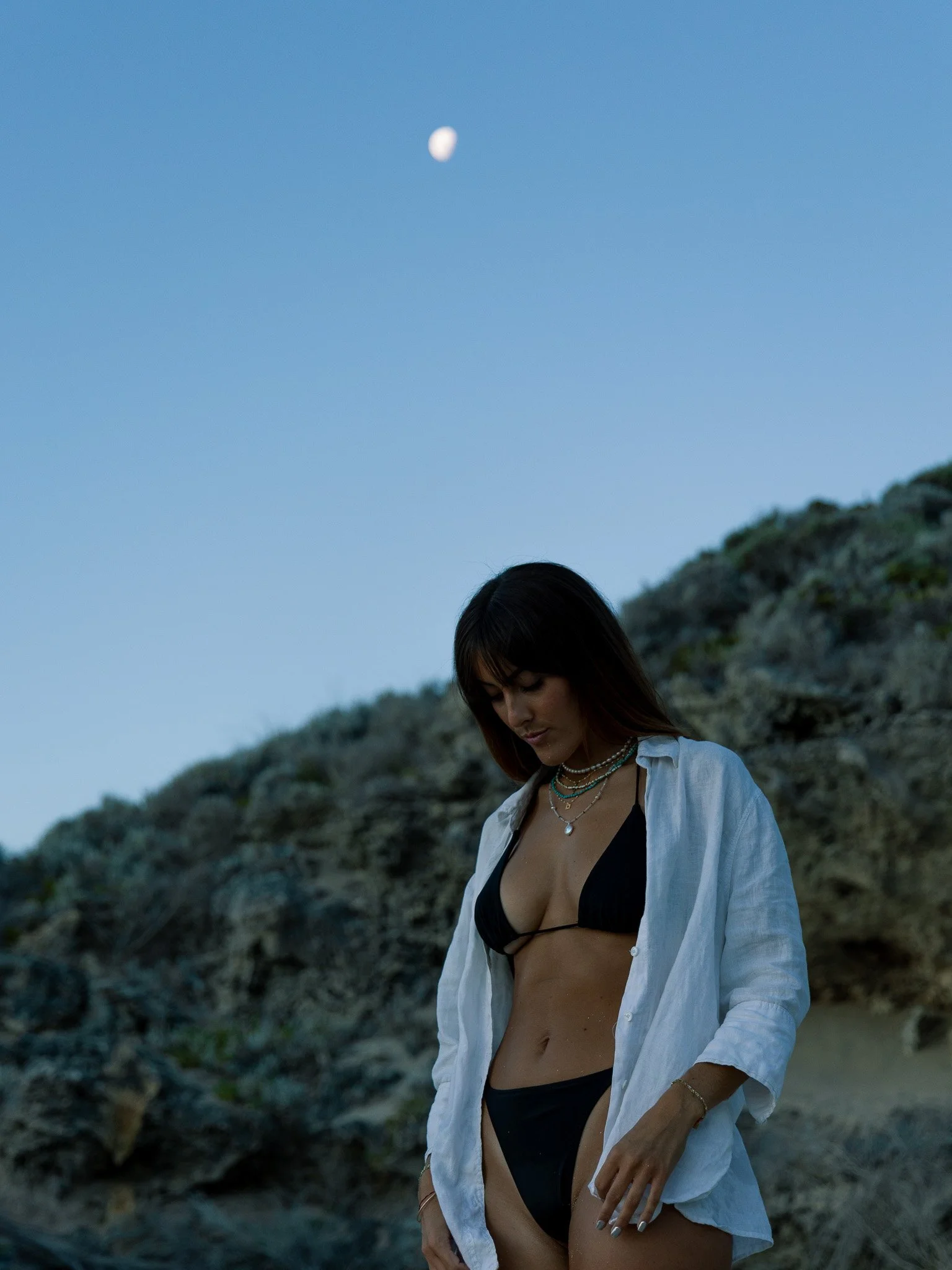 A woman in a black bikini with a white open shirt stands outdoors near rocks during dusk, with the moon visible in the sky overhead.