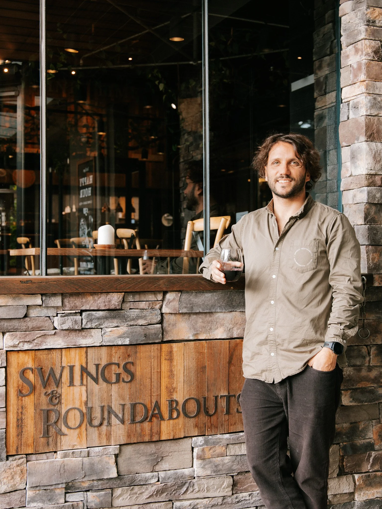 A man with curly hair and a beard standing outside a restaurant called 'Swings & Roundabouts,' holding a glass of red wine, dressed in a beige button-up shirt and dark pants, with a brick and stone wall behind him.