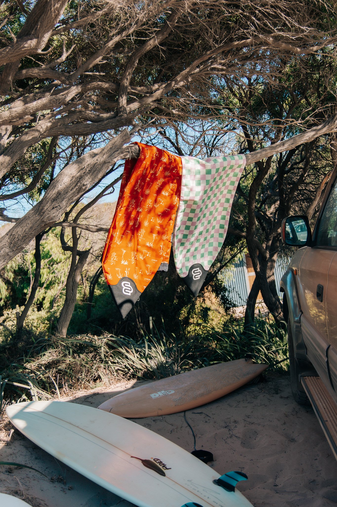 Beach scene with surfboards on sand, orange and patterned fabrics hanging on a tree, and a parked vehicle.
