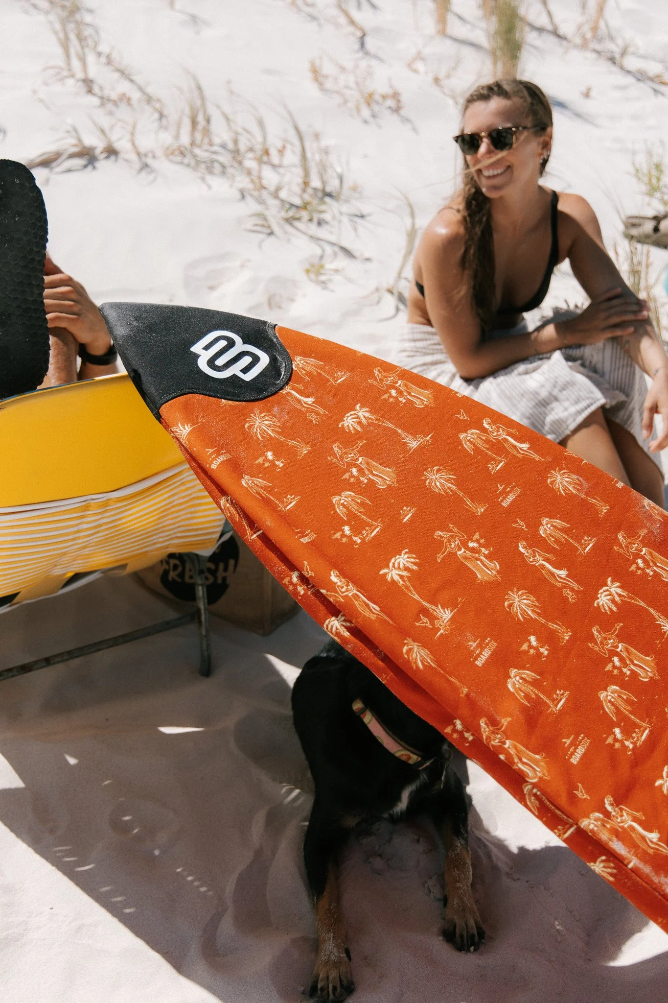 Smiling woman on a beach sitting on a towel, wearing sunglasses and a black bikini top, with a dog under a beach umbrella.