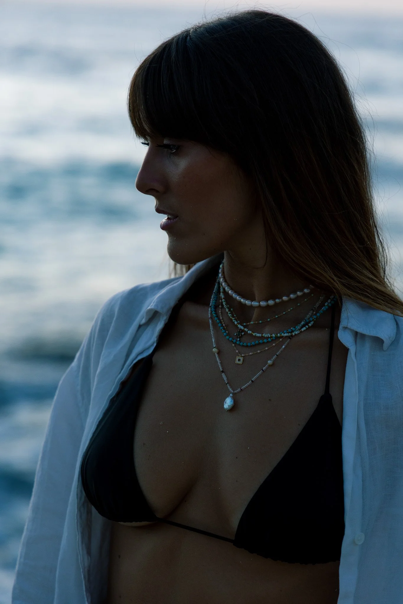 A woman with dark hair wearing layered necklaces, a white shirt open over a black bikini top, standing near the ocean with waves in the background.