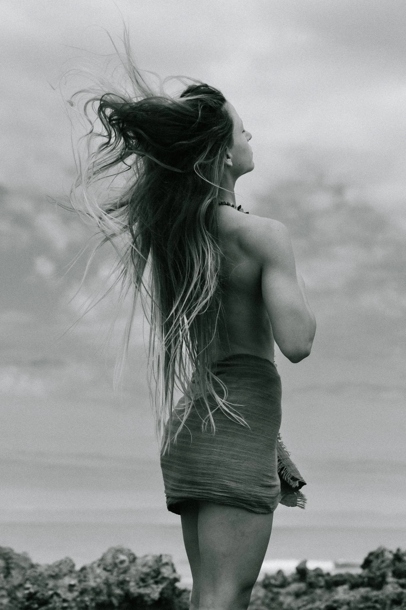 A black and white photo of a woman with long hair blowing in the wind, standing outdoors on a rocky landscape with her arms crossed in front of her, looking to the side.