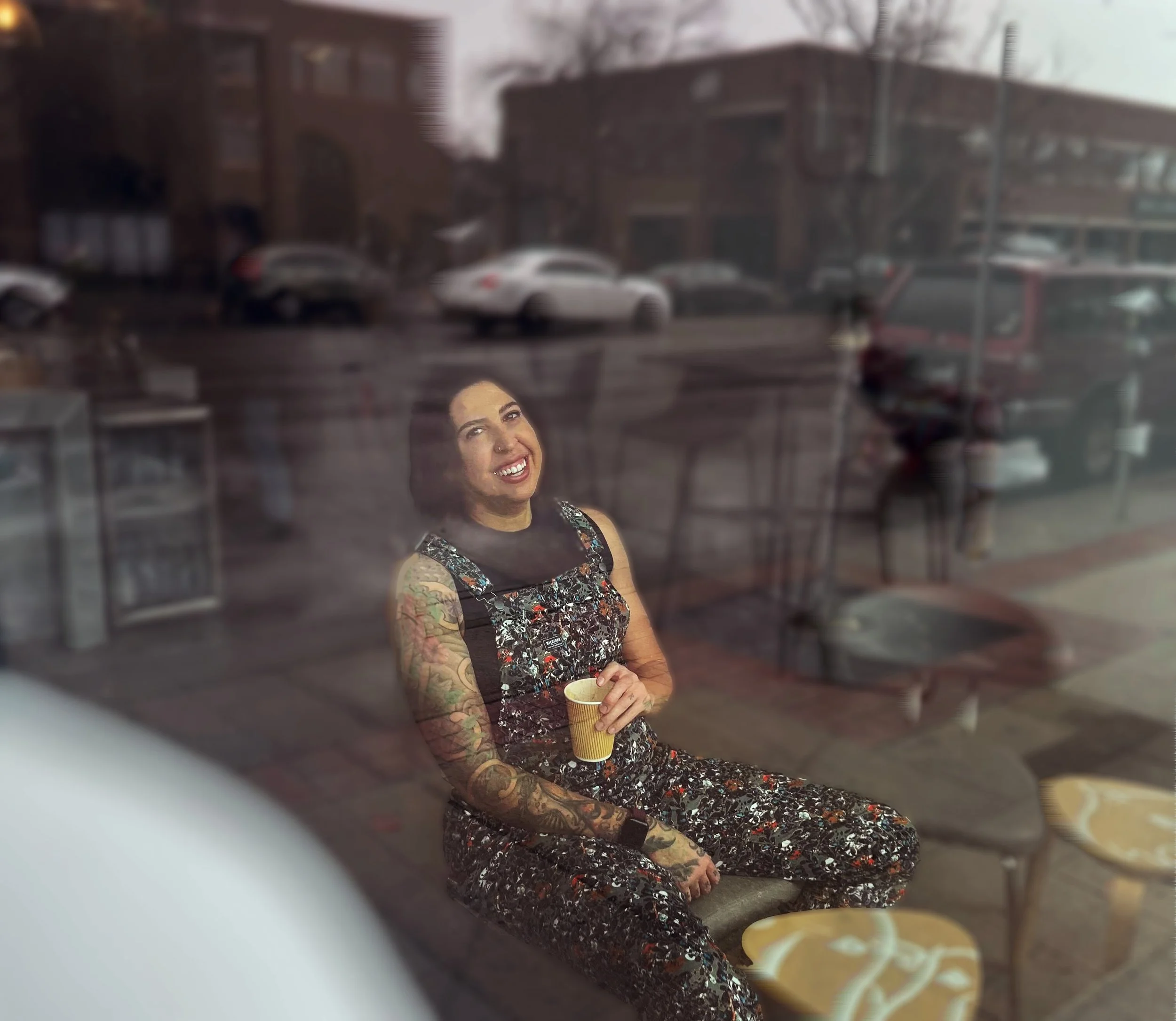 Hallie smiles at the camera through the cafe's window. The reflection of Broadway separates them.