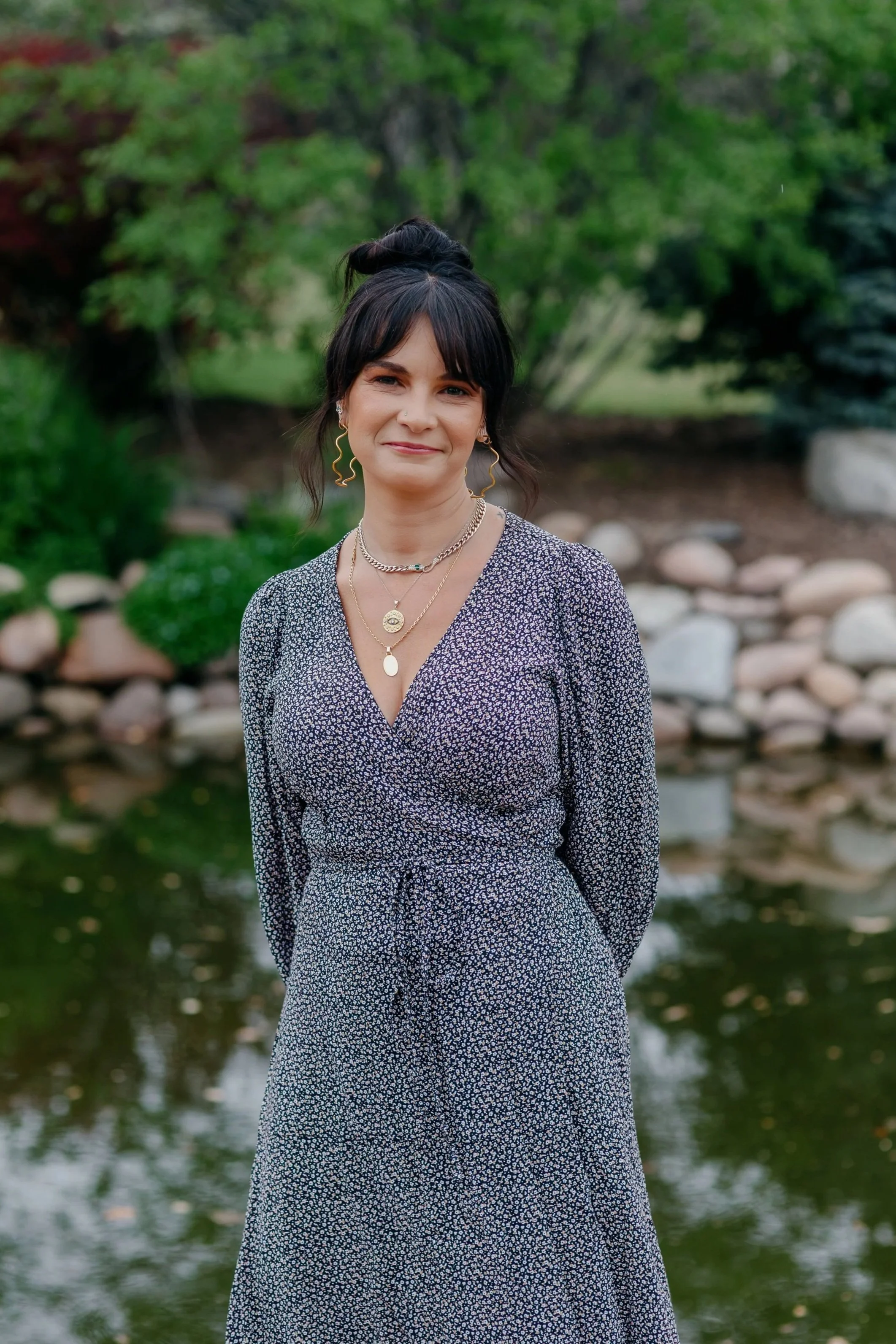 Rene Walter with short black hair styled in a messy bun, wearing large hoop earrings, a nose piercing, a gold necklace, and a dark green ribbed top, outdoors in a bright setting.