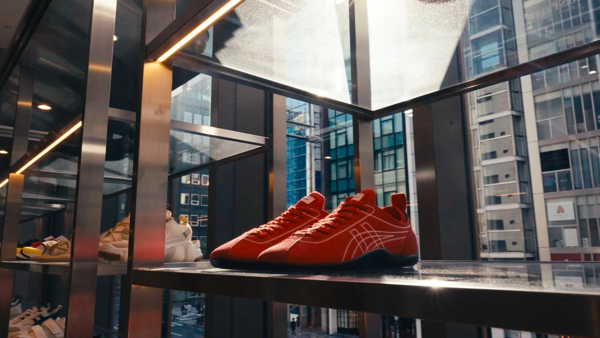 Close-up of red sneakers displayed on a glass shelf inside a modern urban shoe store with large windows and city buildings outside.