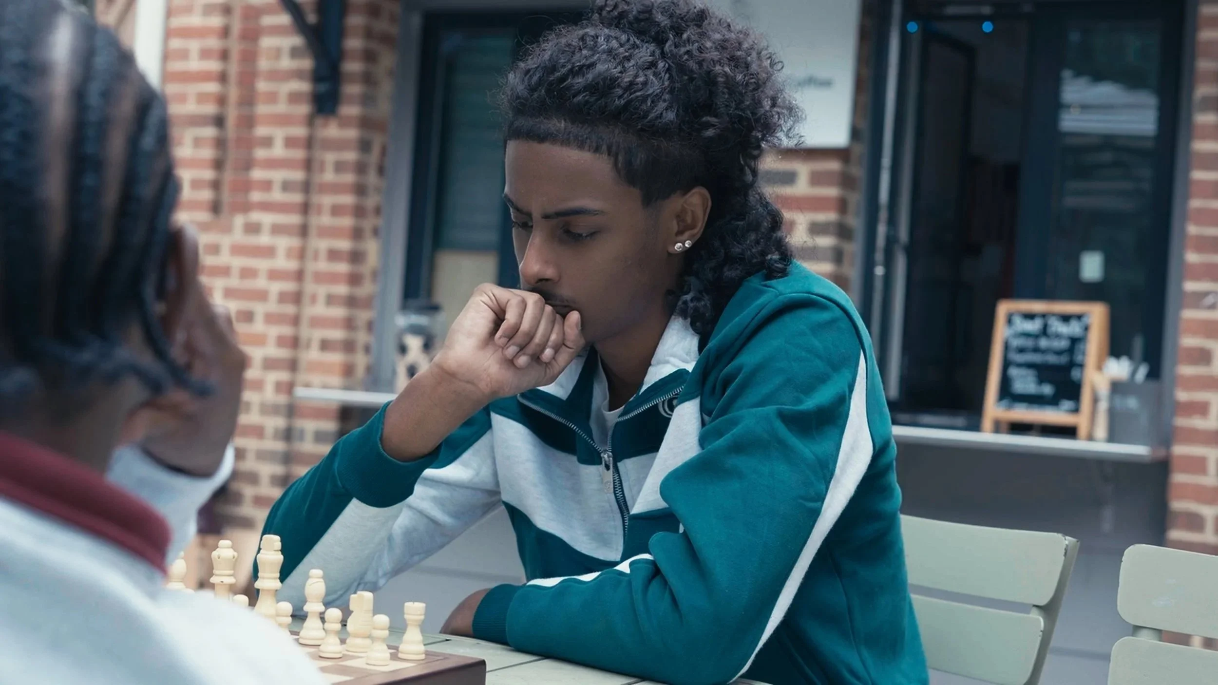Two people playing chess outside, with one person in focus appearing thoughtful, and the other person partially visible in the foreground. The setting is outdoors, near a brick building.