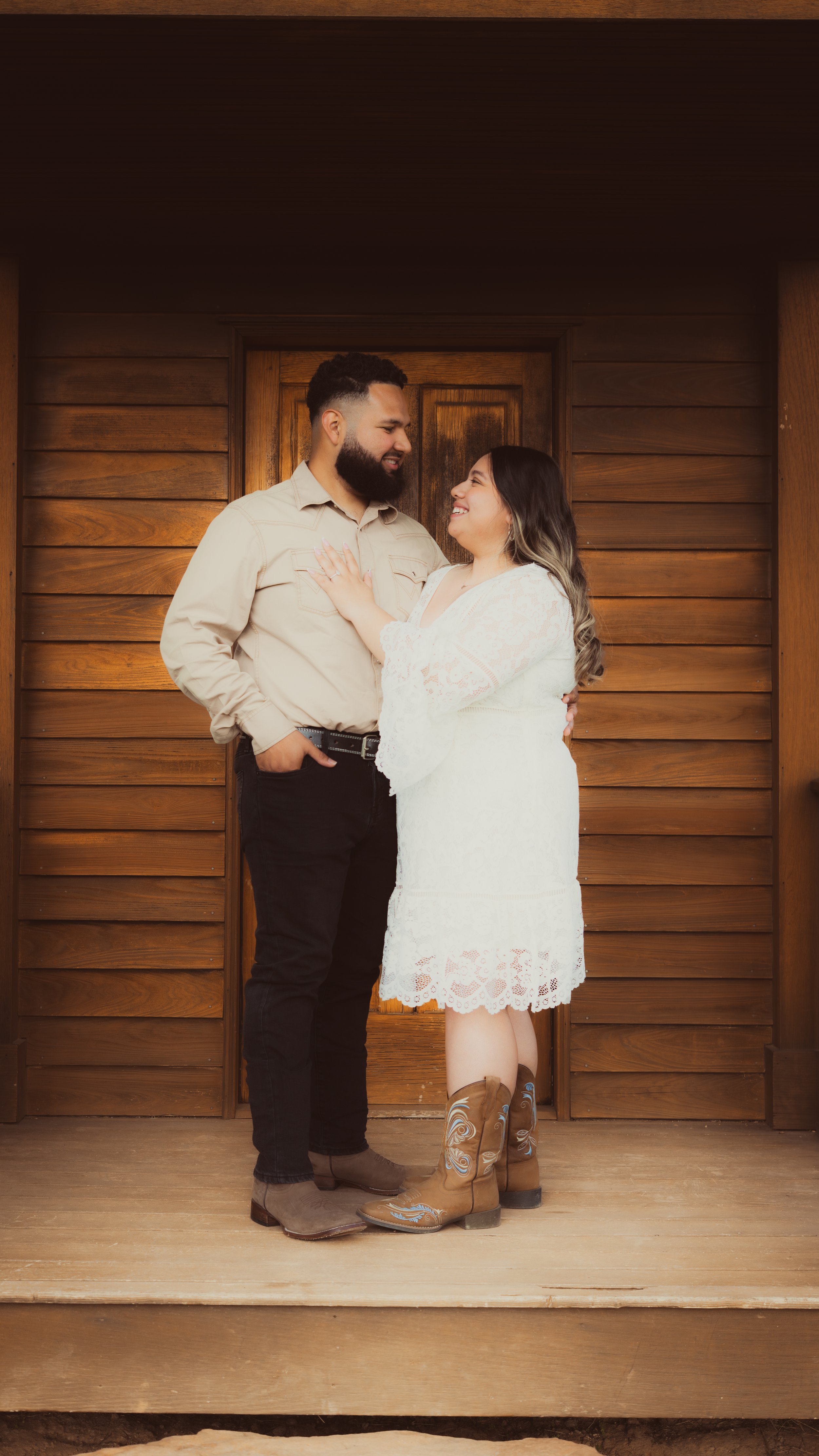 A couple standing close together, looking into each other's eyes, in front of a wooden wall. The woman is wearing a white lace dress and cowboy boots, and the man is wearing a light-colored shirt and black jeans.