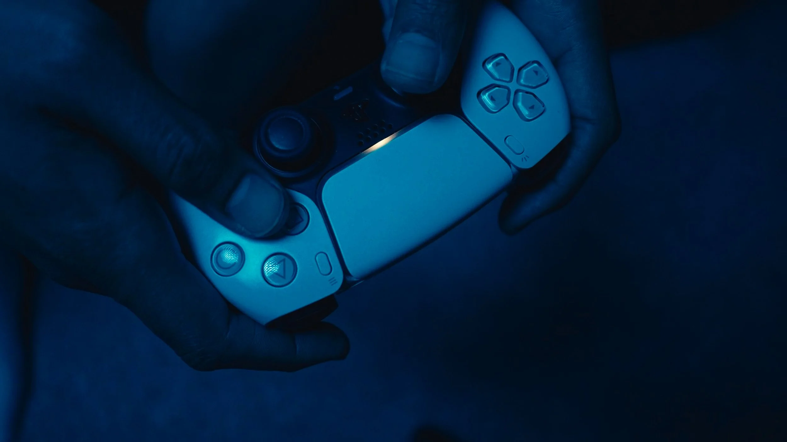 Close-up of a person's hand holding a remote control with illuminated buttons, in a dark environment with blue lighting.