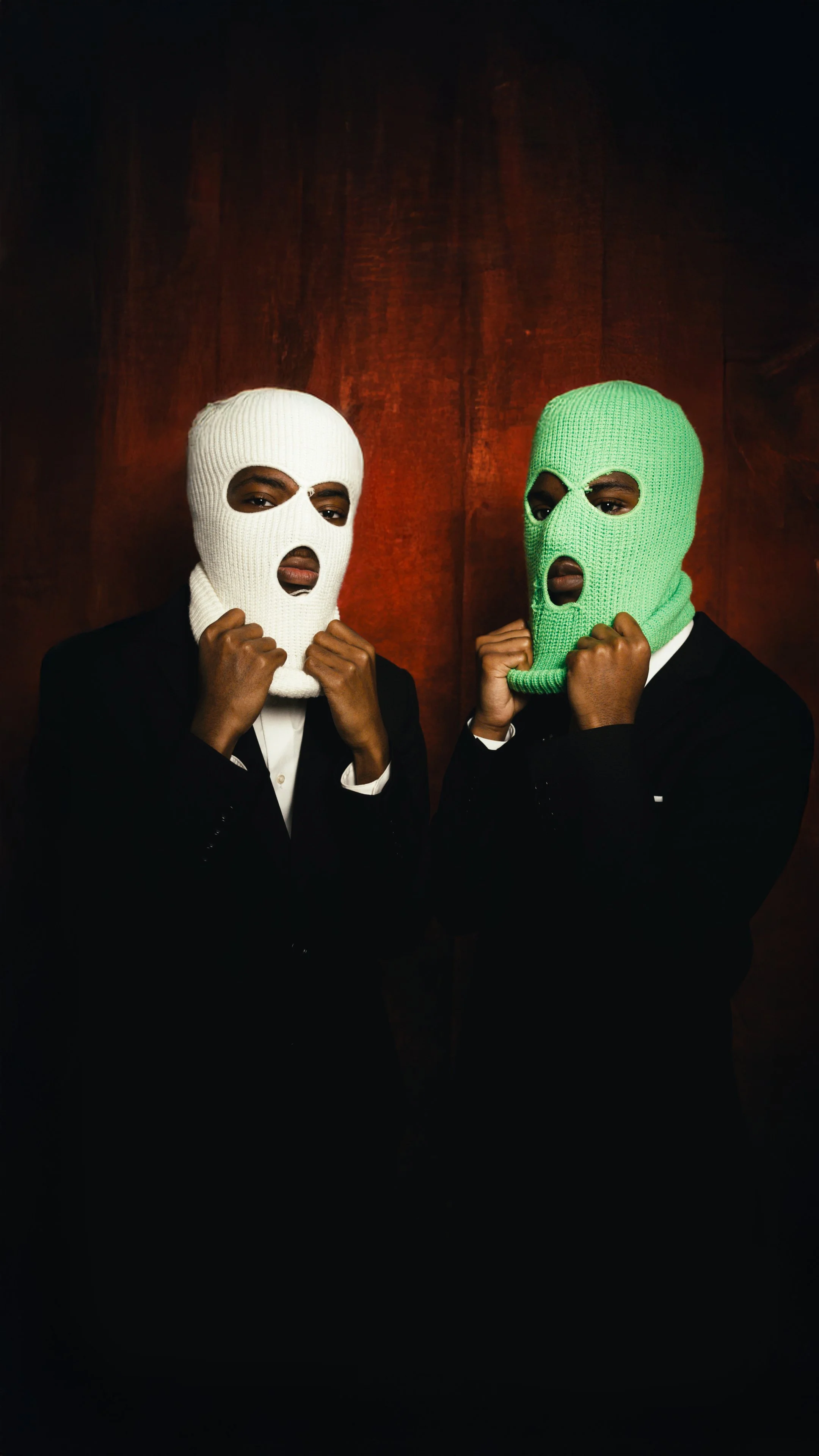 Two men in black suits wearing colored ski masks, one white and one green, standing against a dark red background.
