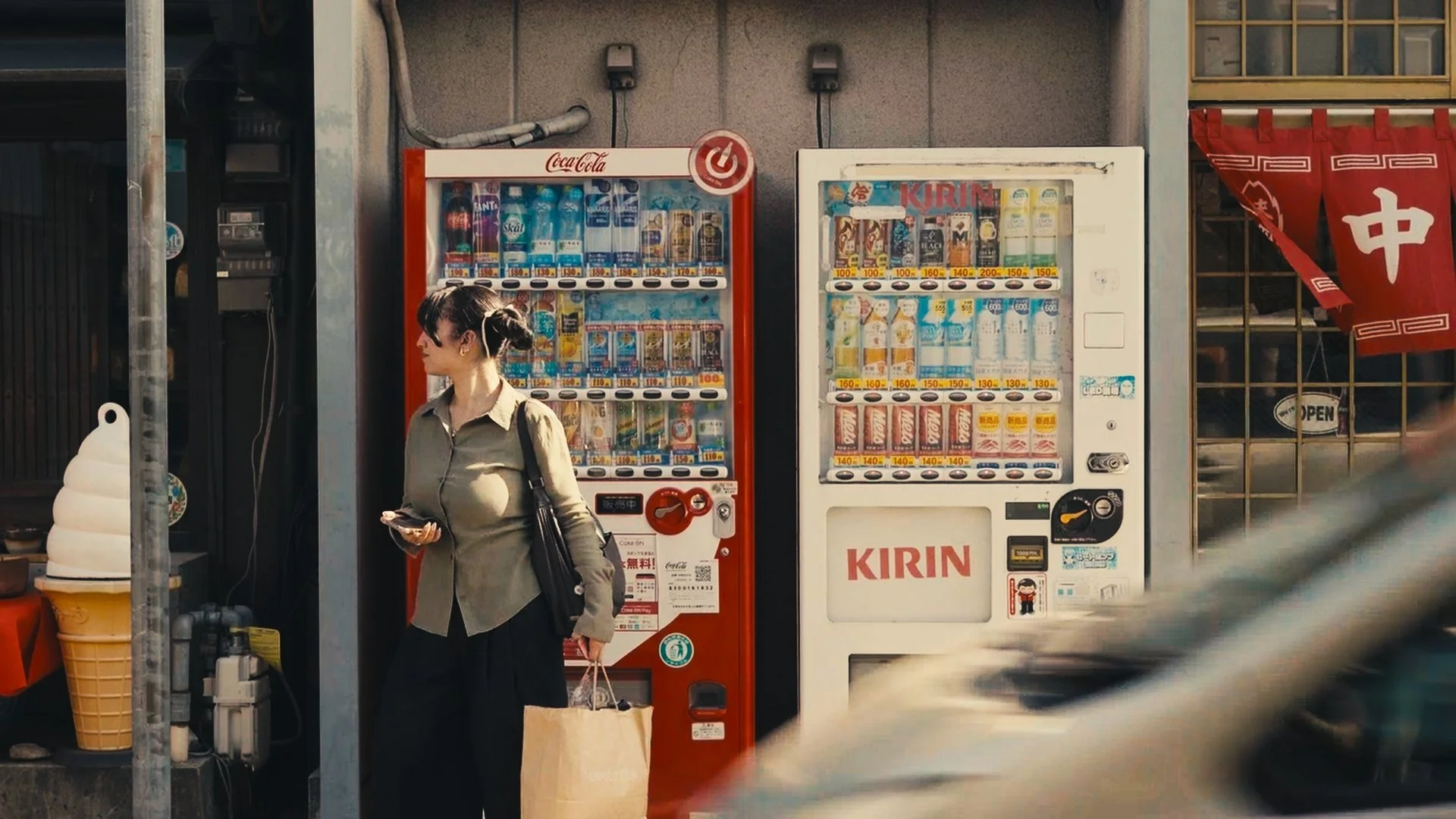 A woman standing in front of two vending machines with beverages, holding a paper bag and a phone, outside a store with a red banner and a glass door.