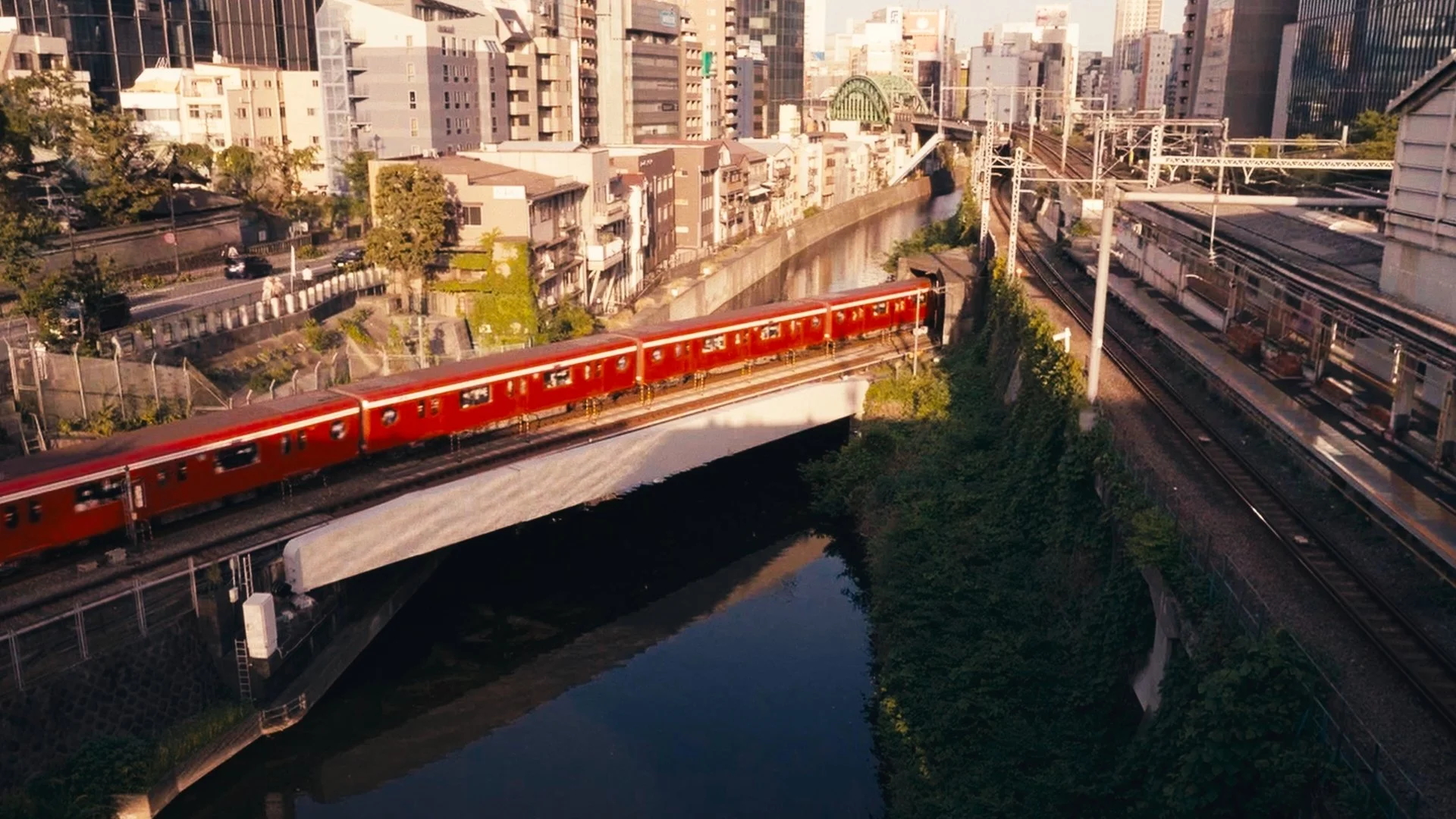 A cityscape with high-rise buildings, a red train crossing a bridge over a river, and train tracks on the right side.