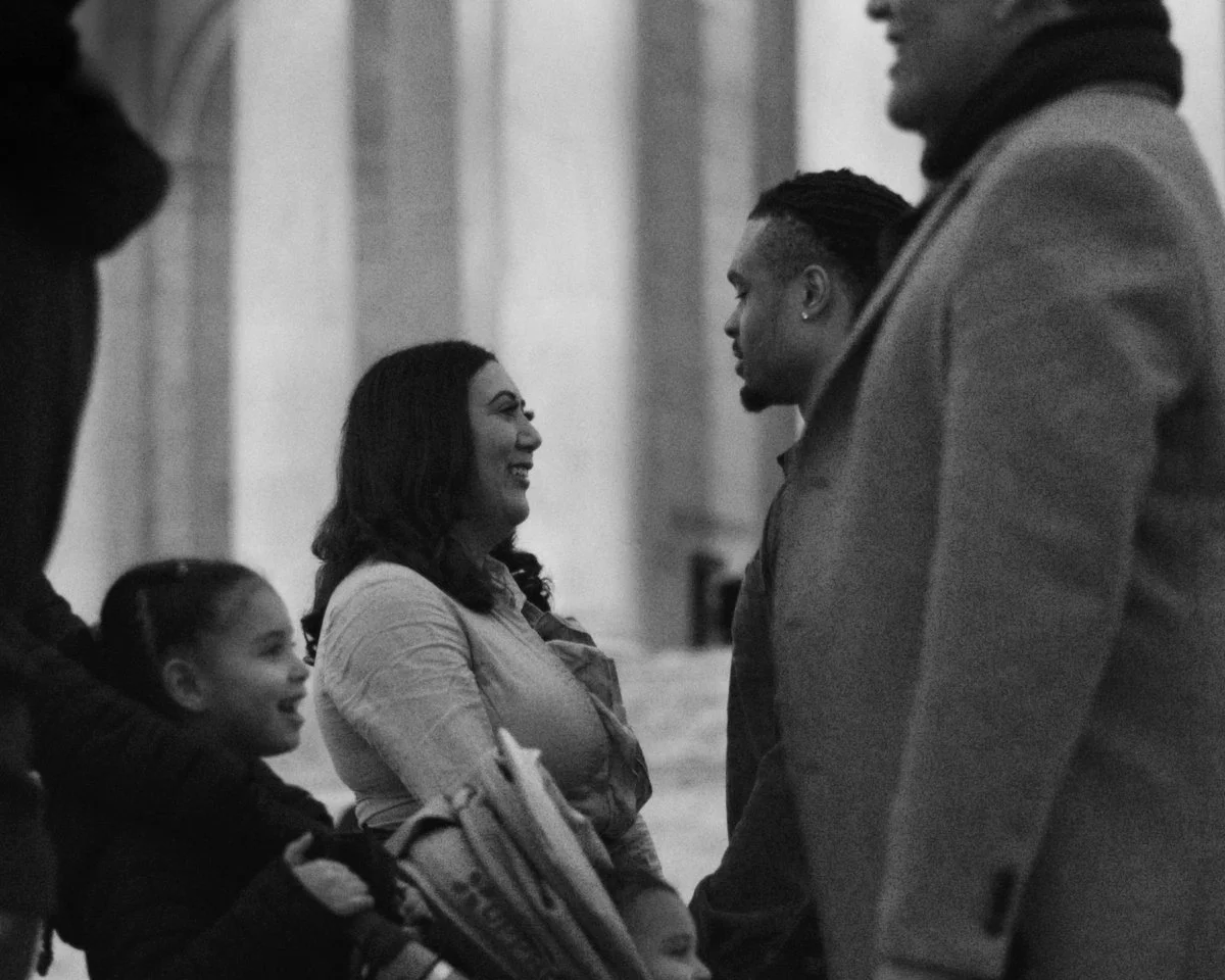 A group of people, including a woman and a man, are engaged in conversation in an indoor setting. The woman is smiling and facing the man, with a young girl standing nearby. The image is in black and white.