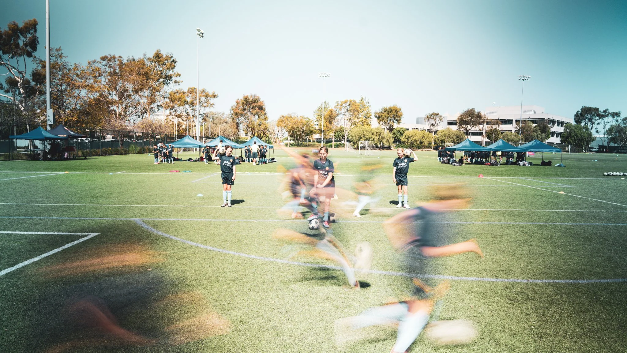 Young girls on a soccer field practicing, with tents and trees in the background, and players running and kicking soccer balls.