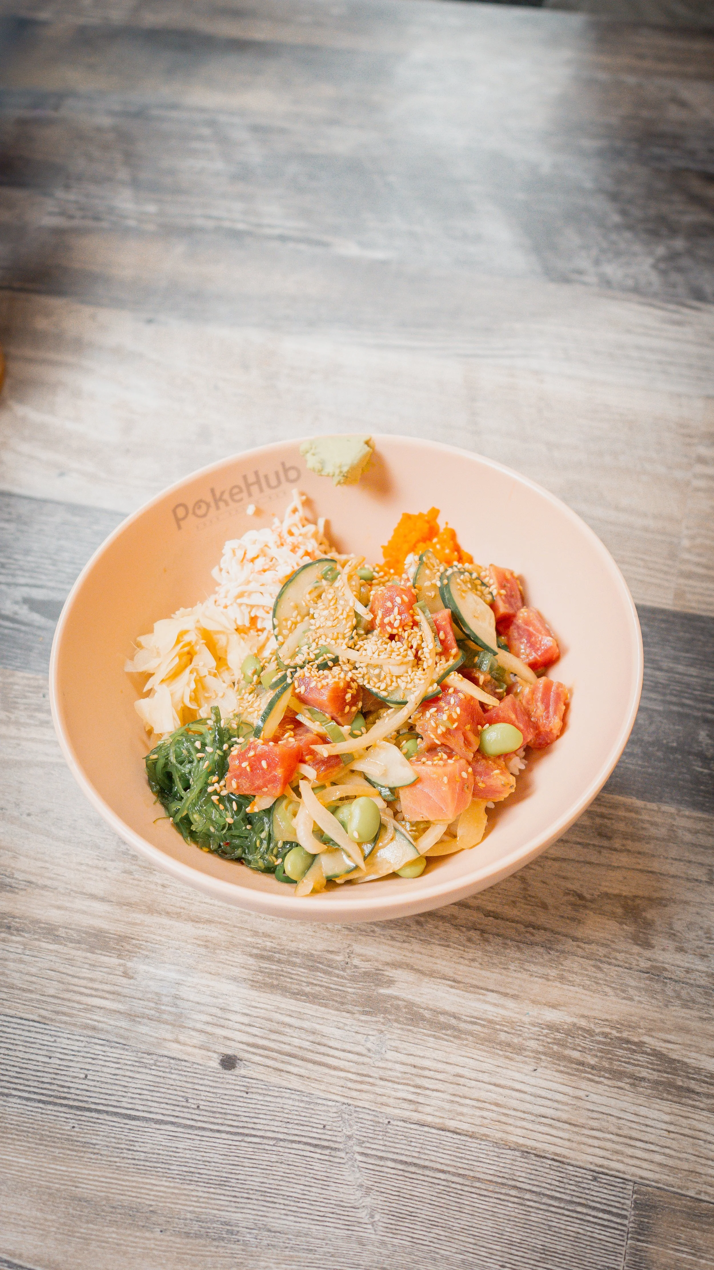 Bowl of poke with cubed raw fish, sliced cucumbers, edamame, shredded vegetables, rice, seaweed, sesame seeds, wasabi, and shredded carrots, on a wooden table.