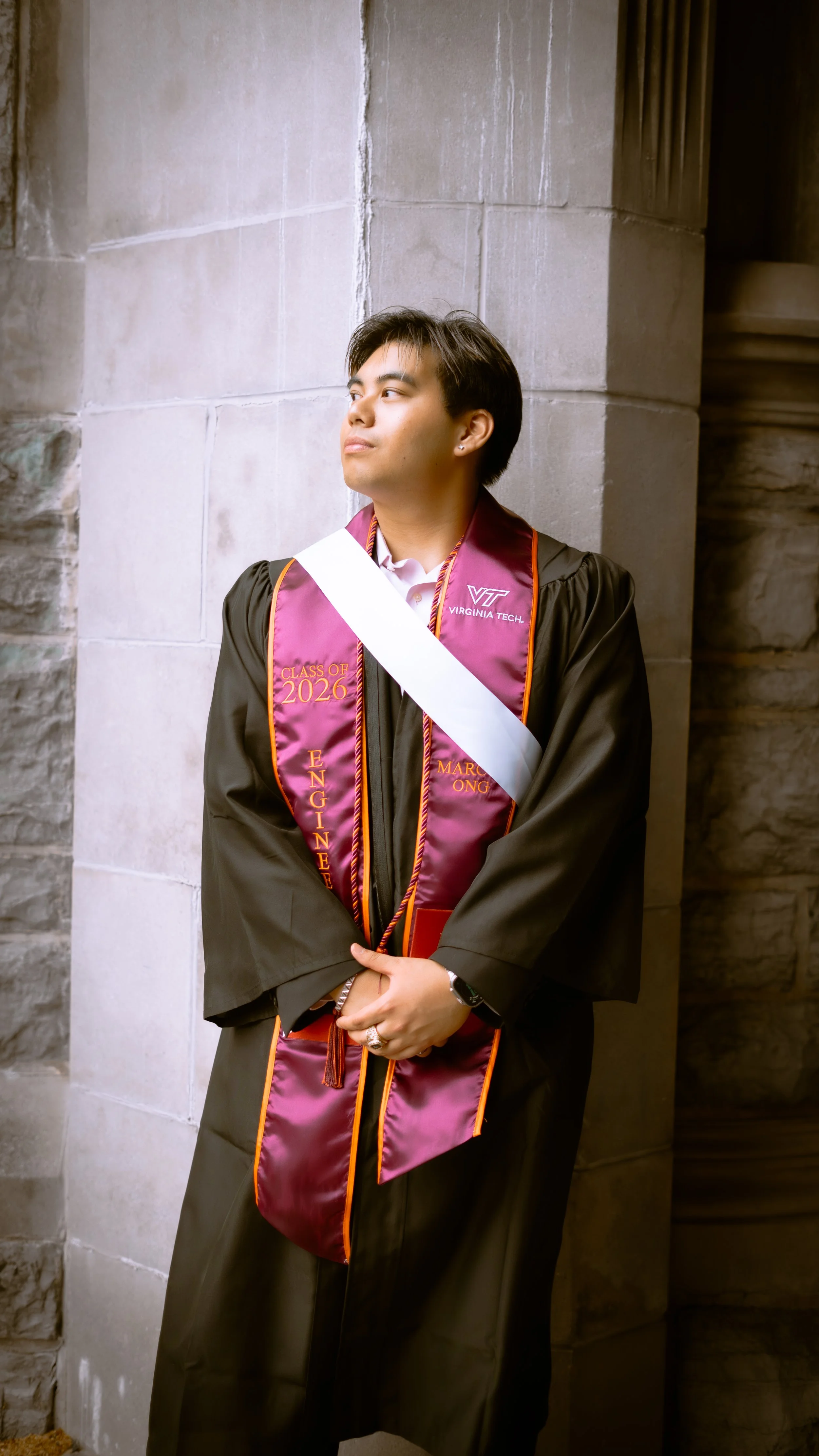 A young man in a graduation gown and cap stands with his arms crossed, looking to the side. He is wearing a maroon and orange stole with 'Class of 2026' and 'Virginia Tech' on it, standing against a stone wall inside a building.